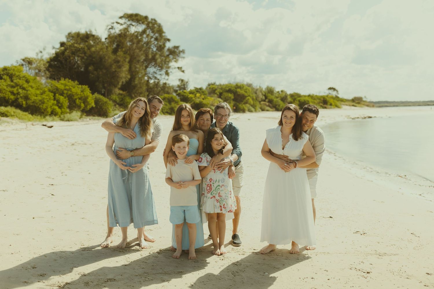 A group in light blue and white dresses walks together on a sunny beach with tropical foliage in the background.