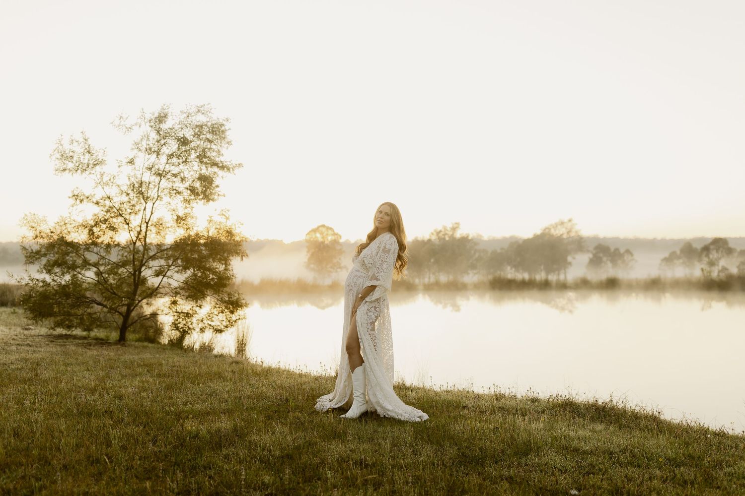 Pregnant woman in white gown standing by misty lake at golden sunrise surrounded by lush green trees.