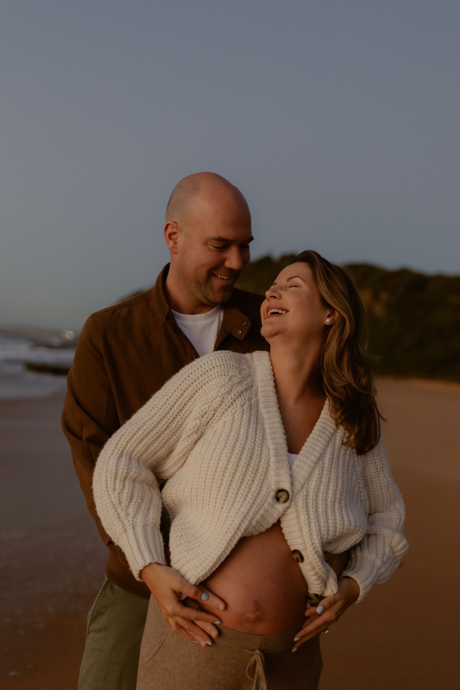 Couple laughing together on beach at sunset, man embracing pregnant womans baby bump tenderly.