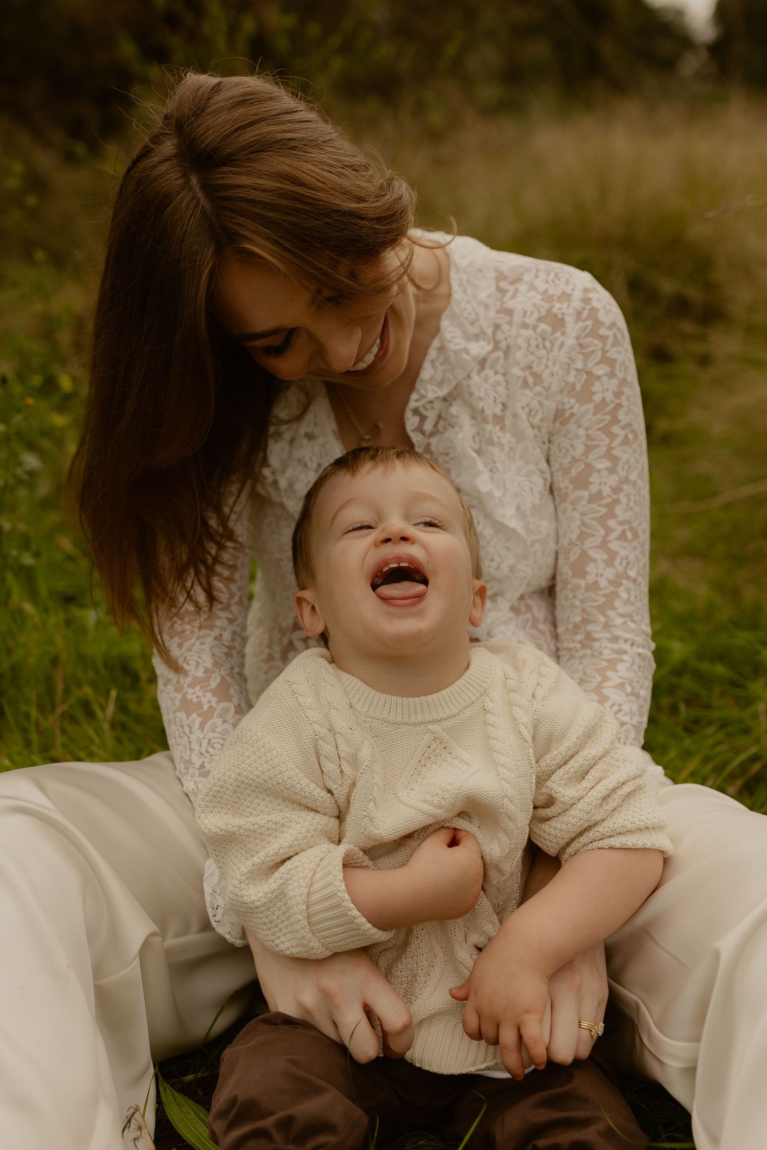 Smiling mother in white lace dress sits in grass with laughing baby boy in cream knit sweater.