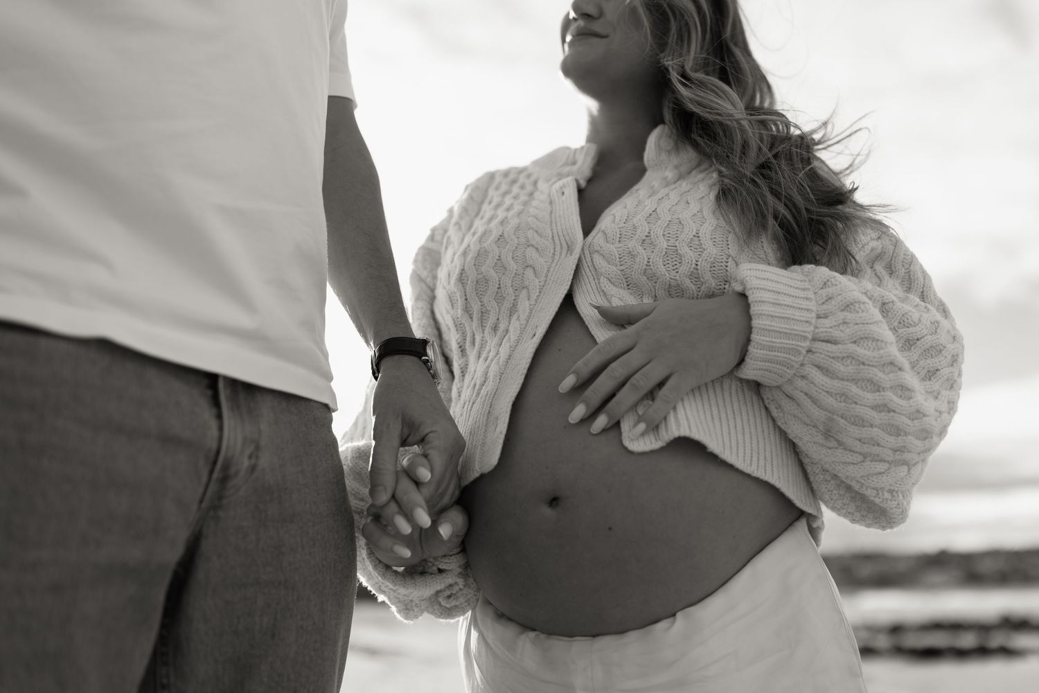 Black and white maternity photo of pregnant woman holding belly, holding hands with partner on beach.