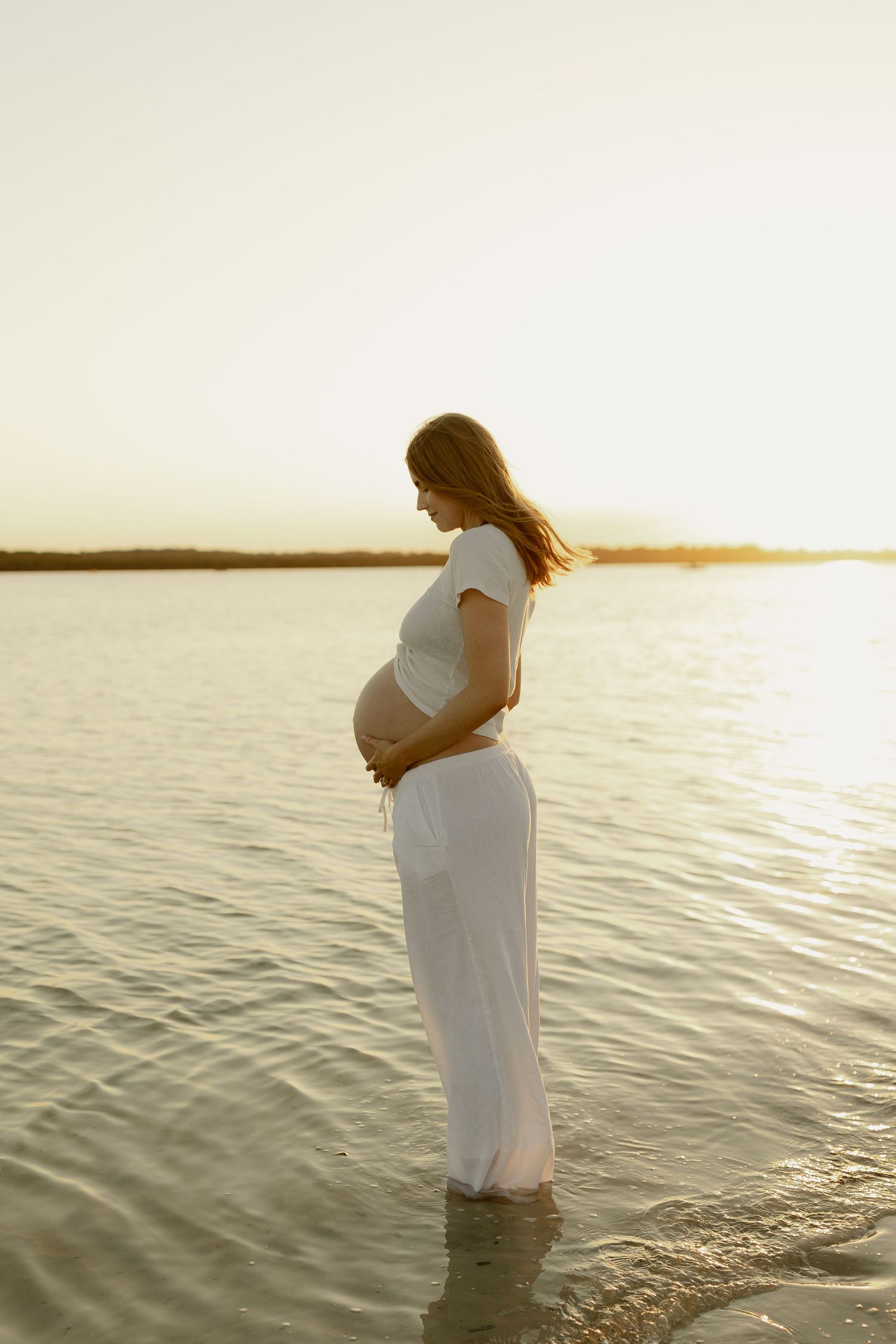 Pregnant woman in white dress standing in shallow water at sunset, cradling her baby bump.