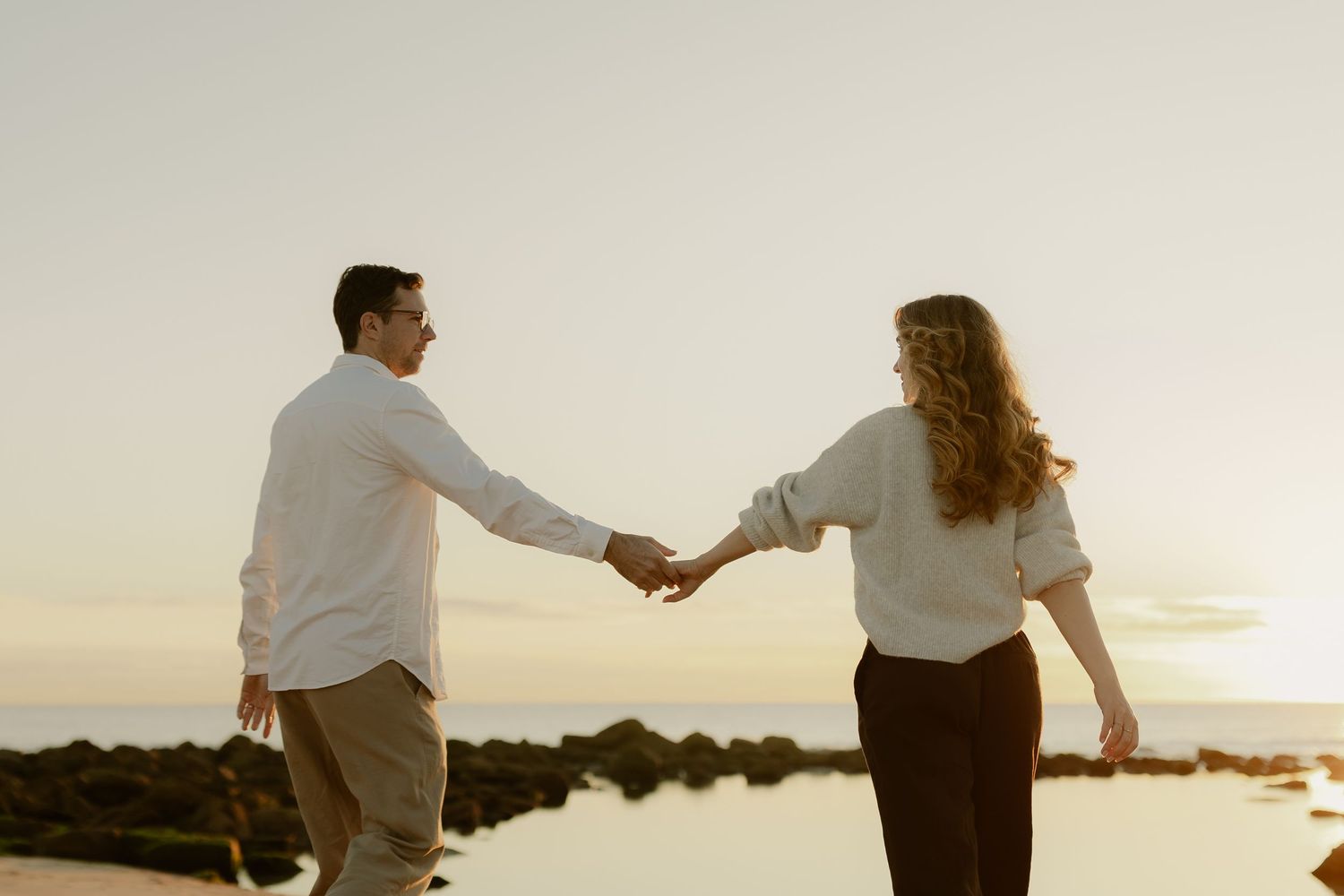 Couple holding hands on a rocky beach at sunset, facing each other in casual white outfits.