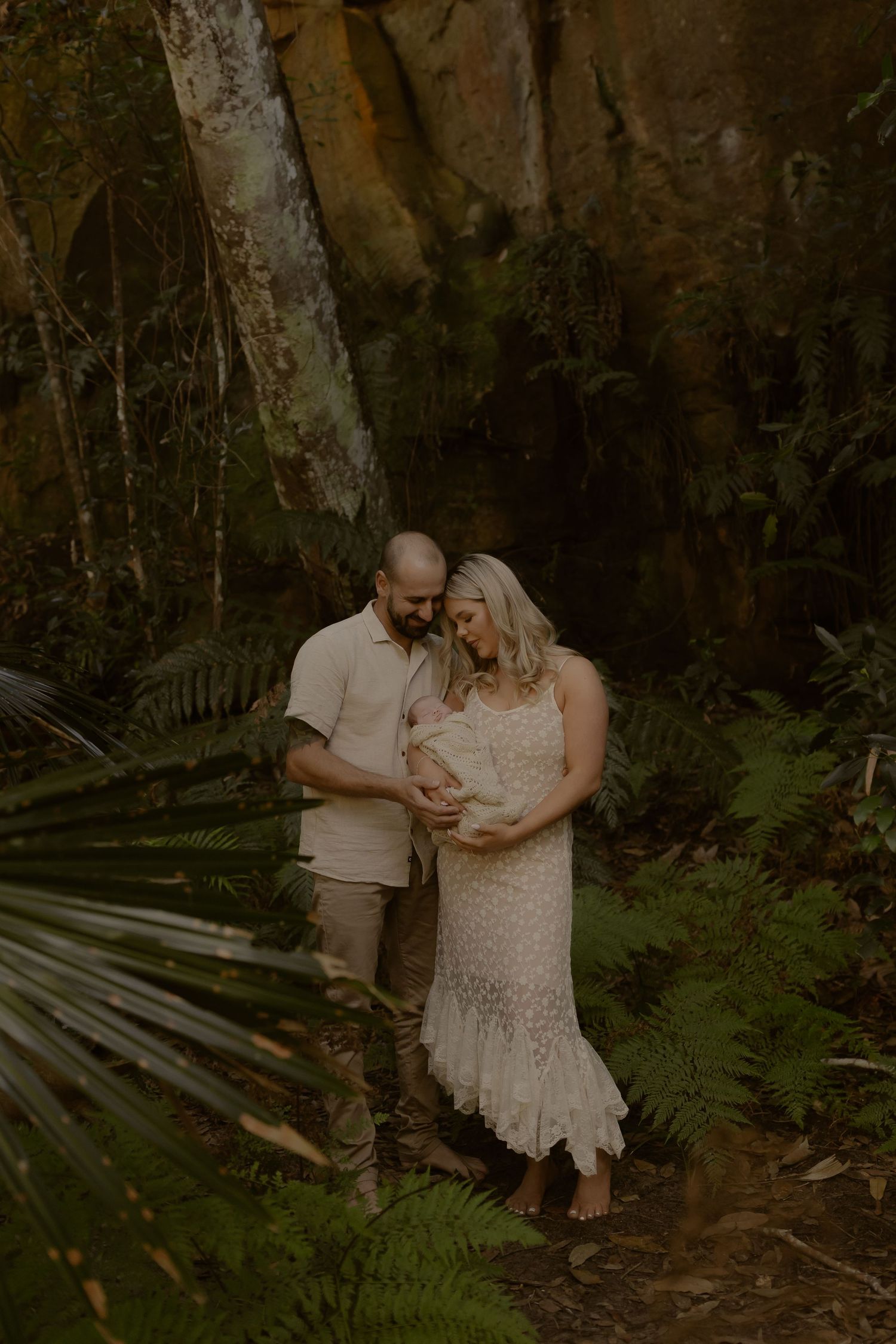 A couple stands in a lush fern forest, lovingly gazing at their newborn baby wrapped in a cream lace blanket.