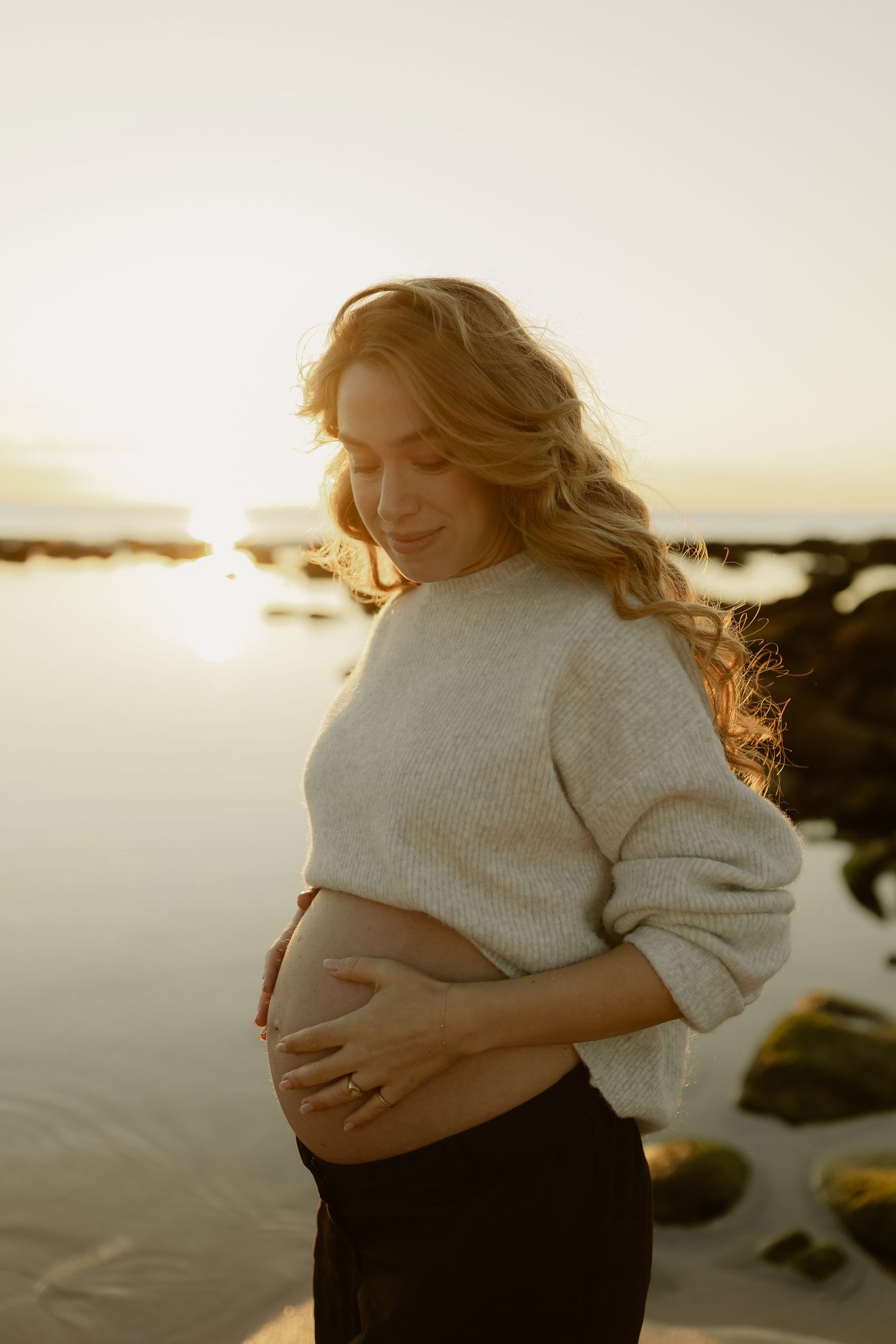 Pregnant woman in cream sweater cradles baby bump on beach at golden hour sunset.