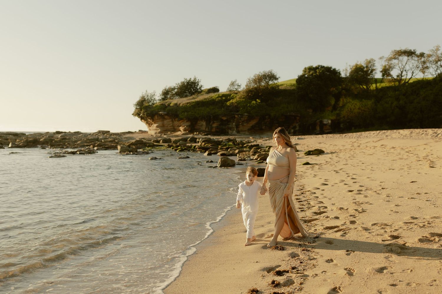 A mother and child walk hand-in-hand along a sandy beach at golden hour, waves gently lapping the shore.