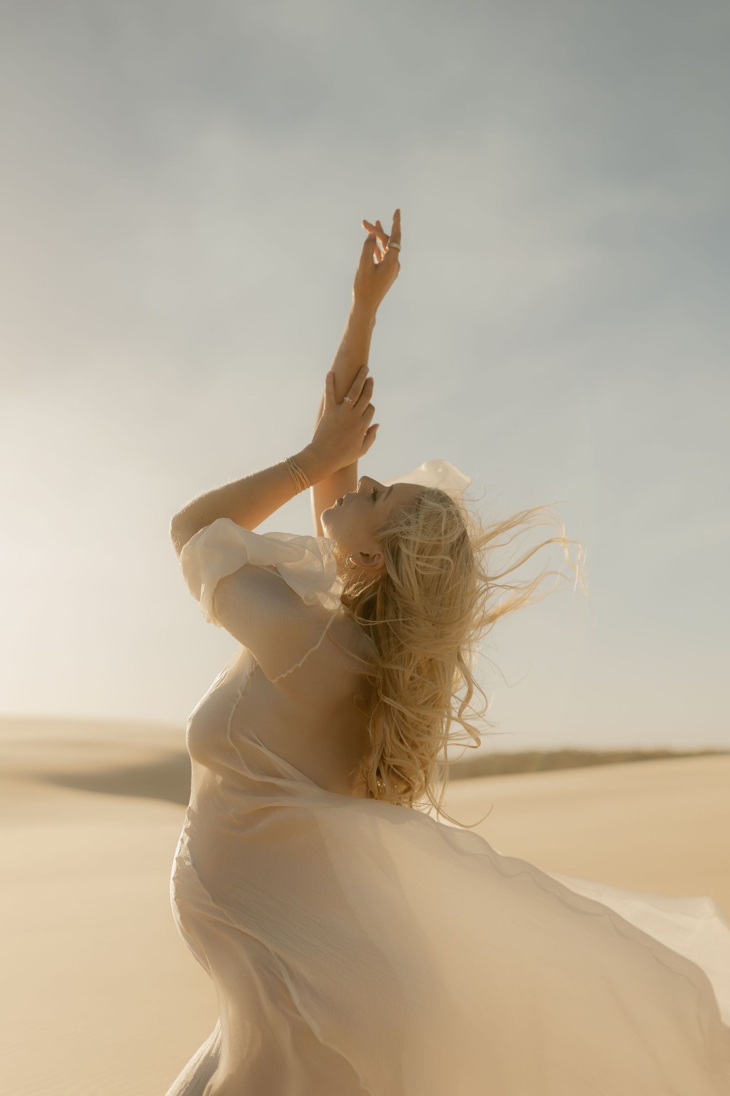 A blonde woman in a flowing white dress reaches arms upward gracefully in a sunlit desert landscape.