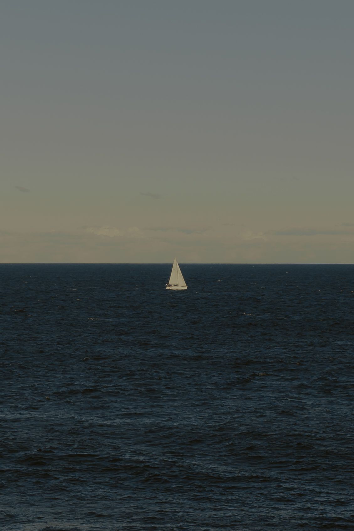 A lone white sailboat navigates calm dark blue ocean waters under an overcast sky at dusk.