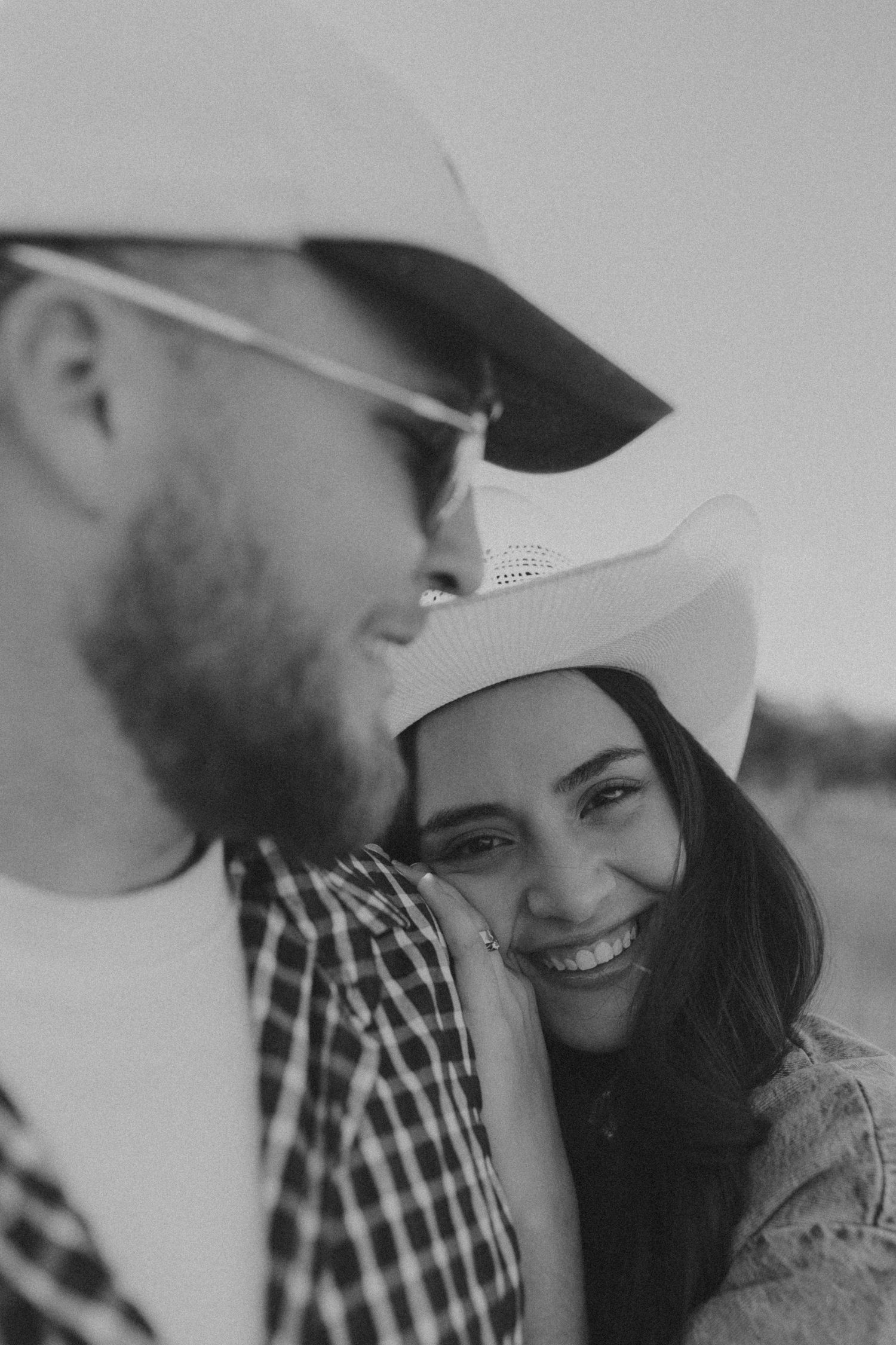 Black and white photo of a smiling woman in cowboy hat leaning on a bearded man wearing sunglasses outdoors.