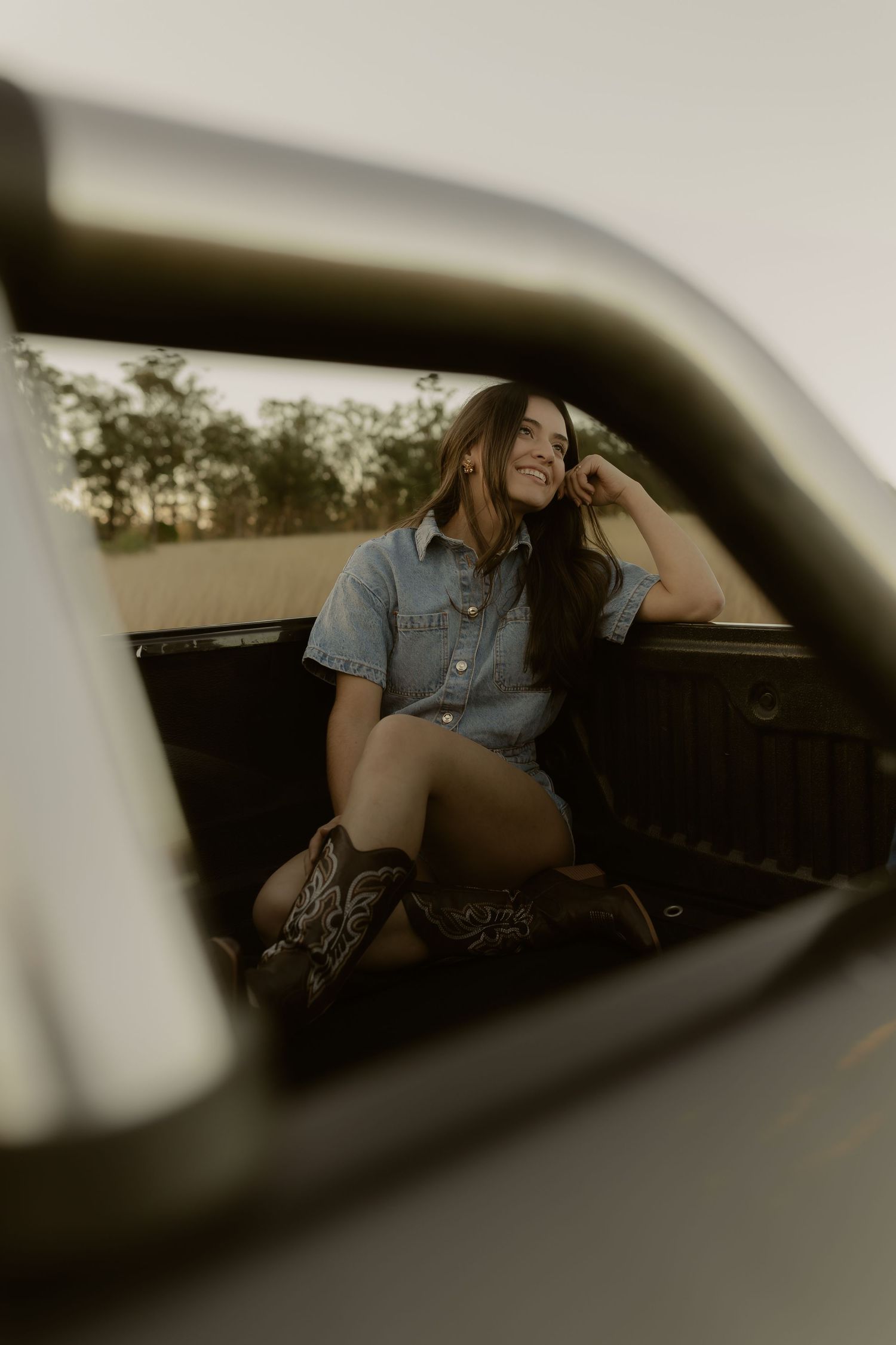 Young woman in denim romper and cowboy boots sitting in truck bed, smiling outdoors.