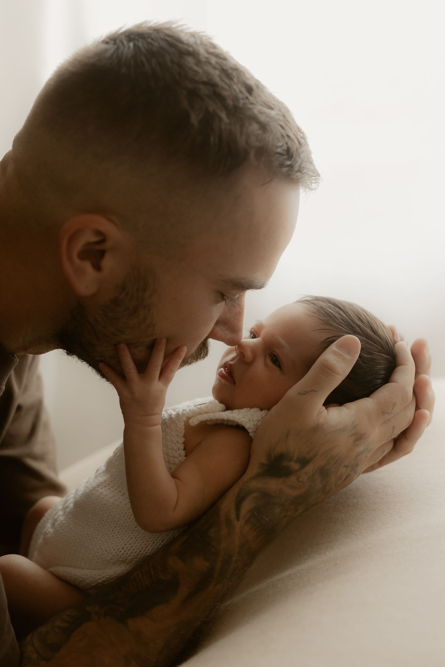 Tattooed father gently touches noses with his newborn baby, cradling the infant tenderly in a soft, warm light.