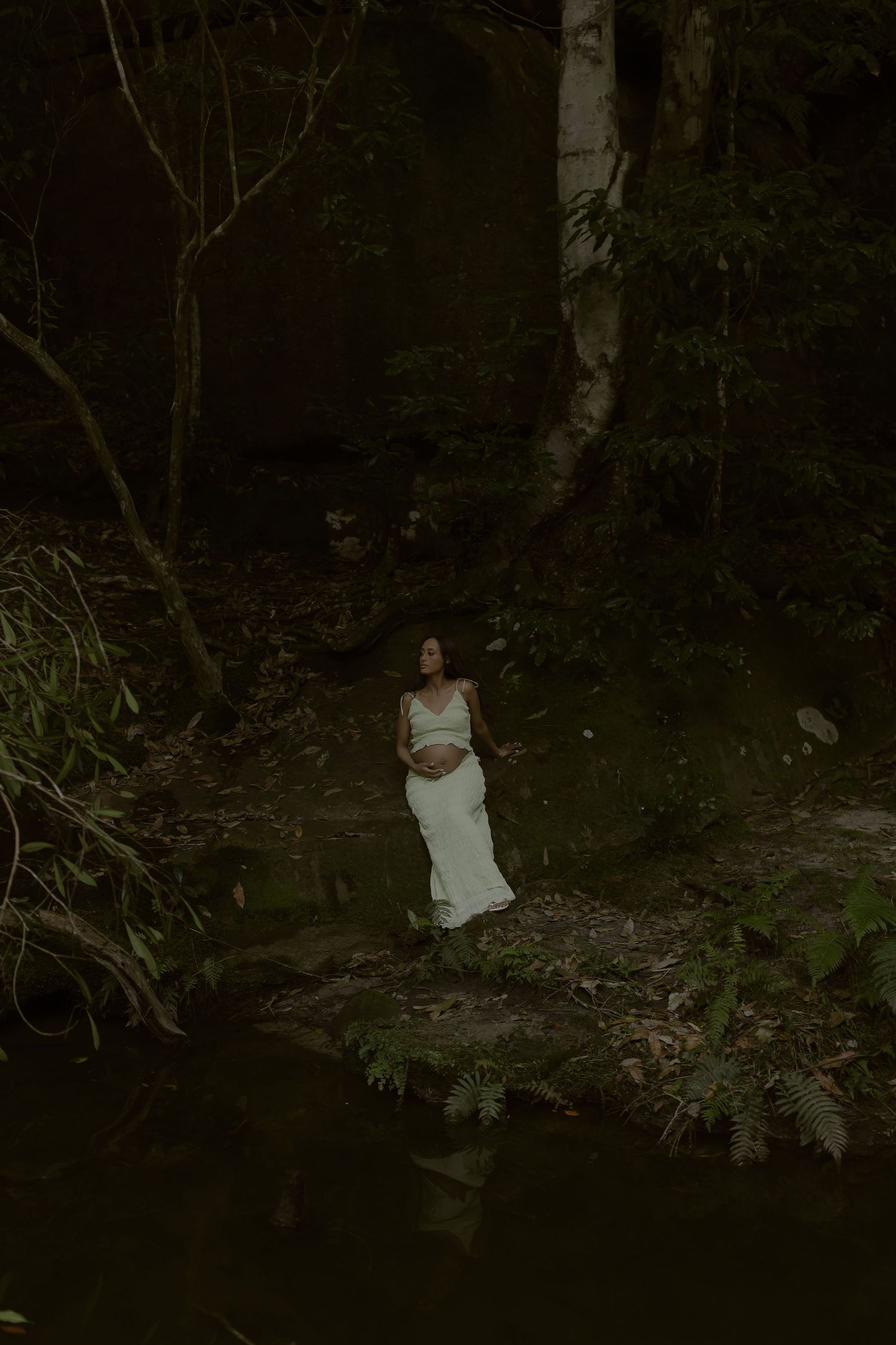 Woman in white dress standing on rocks by a dark forest stream surrounded by lush green foliage.