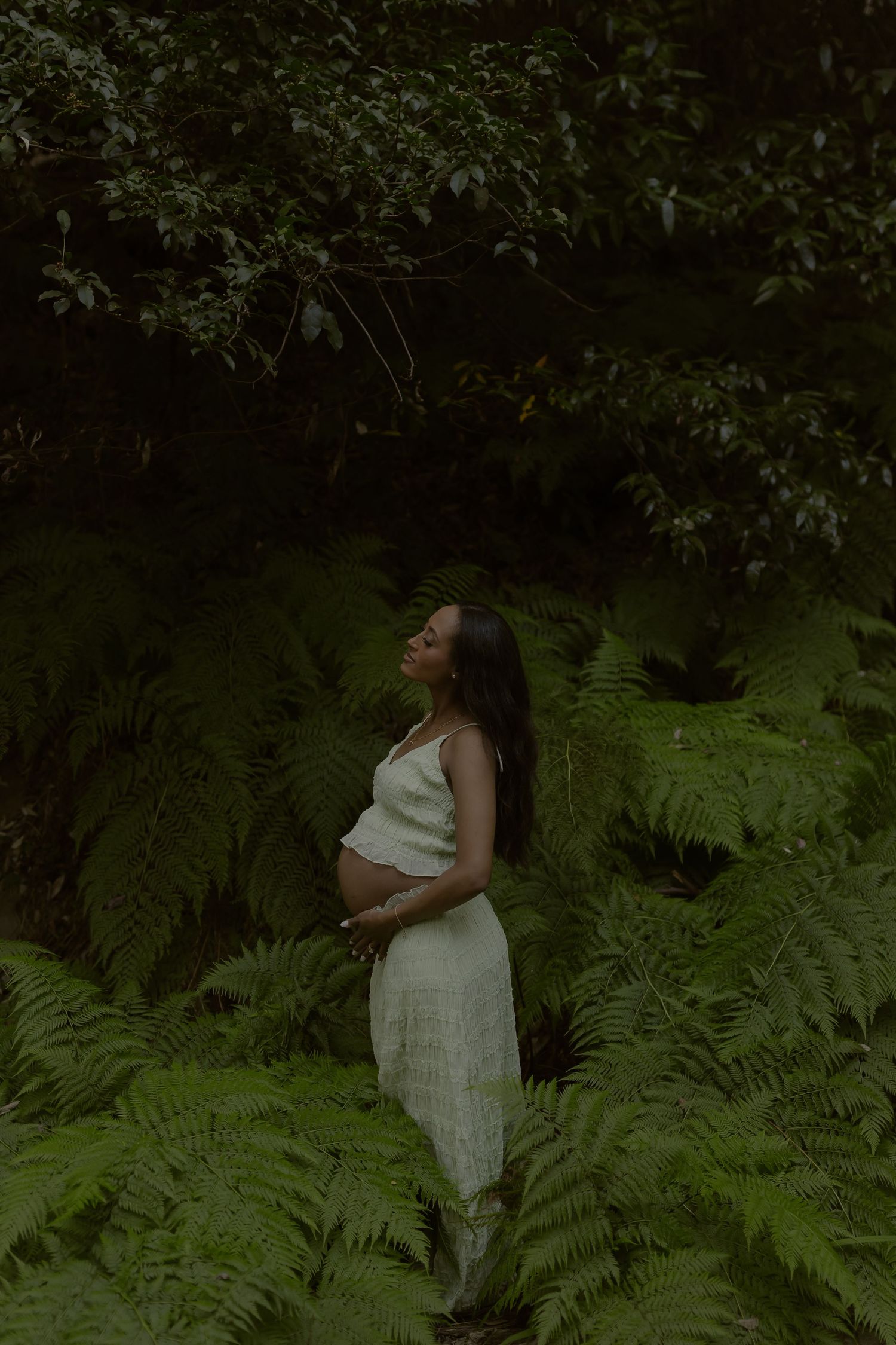 Pregnant woman in white dress standing among lush green ferns in a forest, cradling her baby bump.