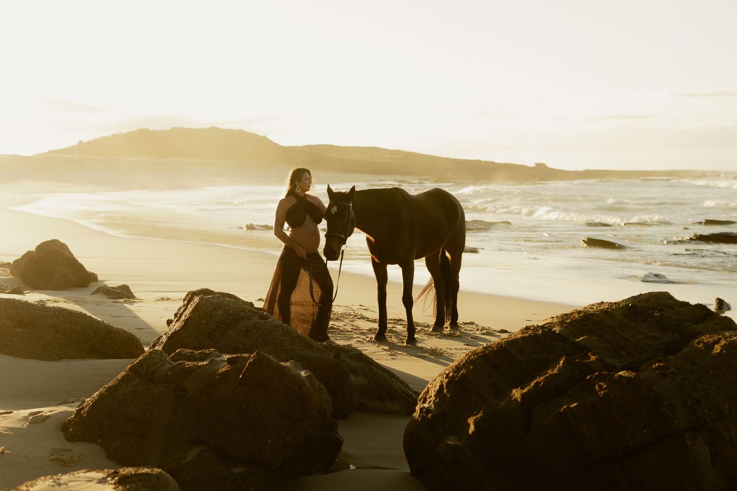 A woman stands with a dark horse on a sandy beach at golden sunset, surrounded by rocks and ocean waves.