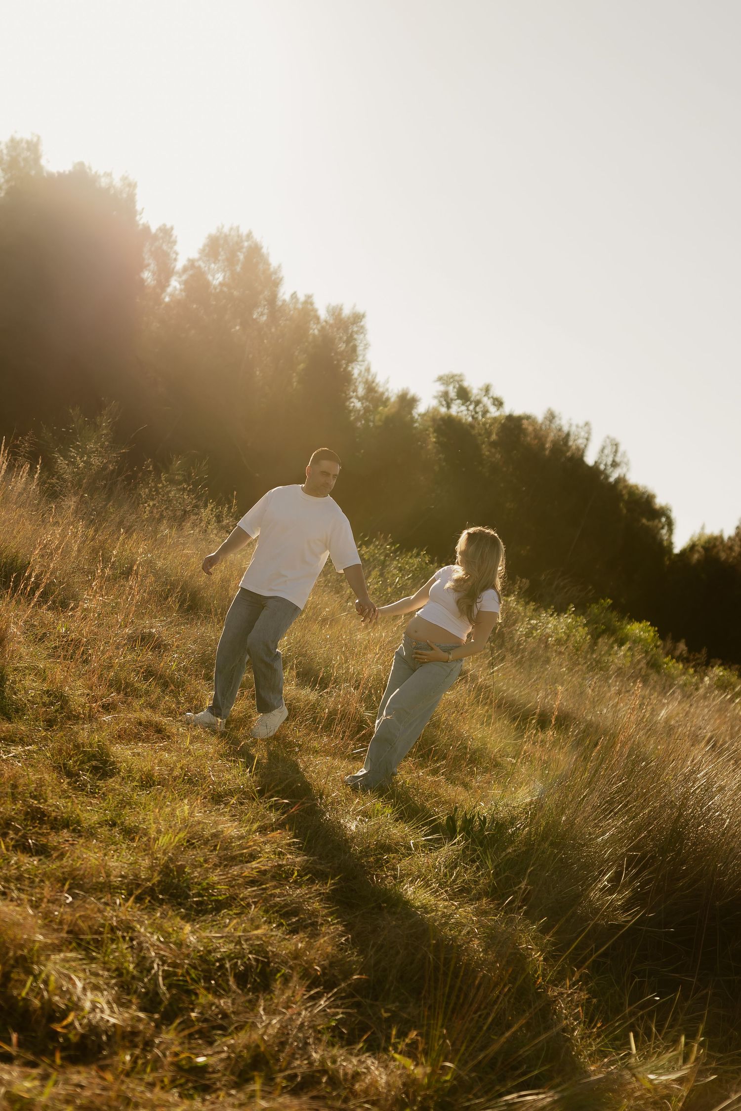 Couple holding hands walking up a sunny grassy hill, wearing white t-shirts and jeans in golden hour light.