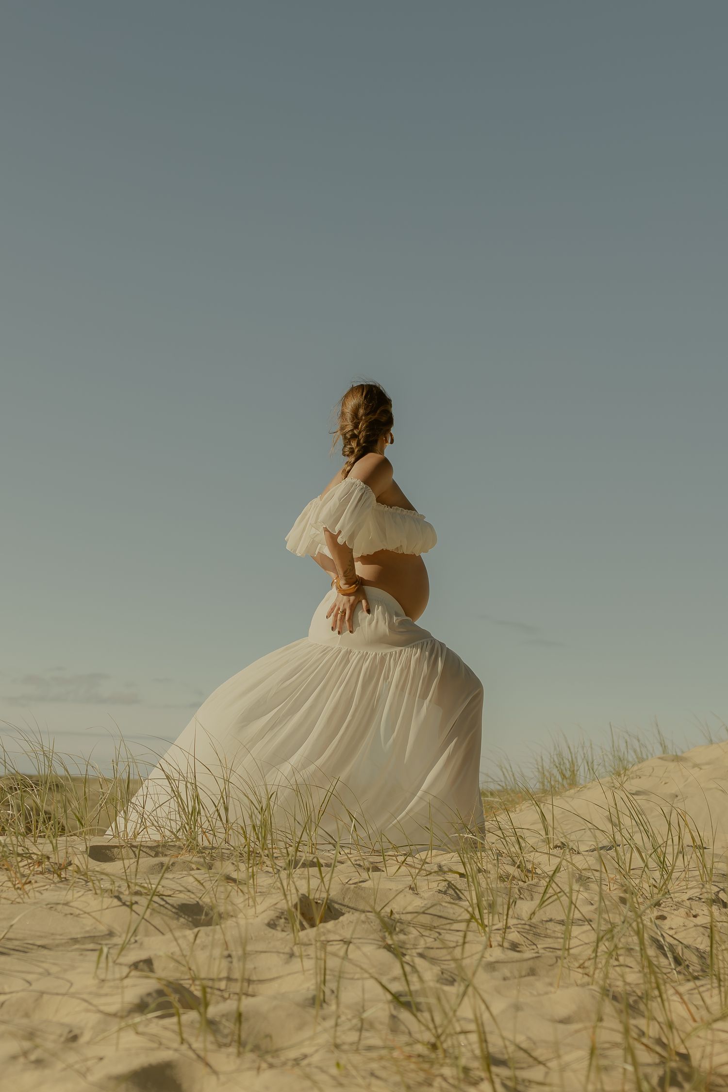 Pregnant woman in elegant white dress stands atop sandy dunes with beach grass under blue sky.