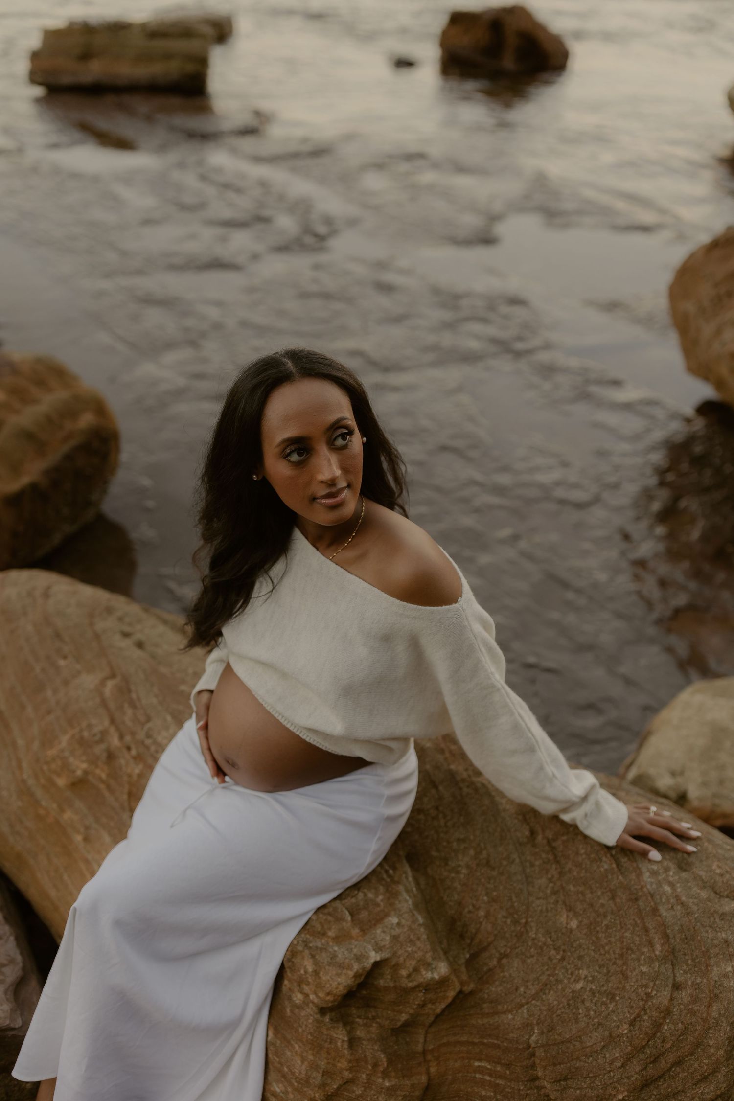 Pregnant woman in off-shoulder white knit top and skirt sitting on rocks by the ocean.