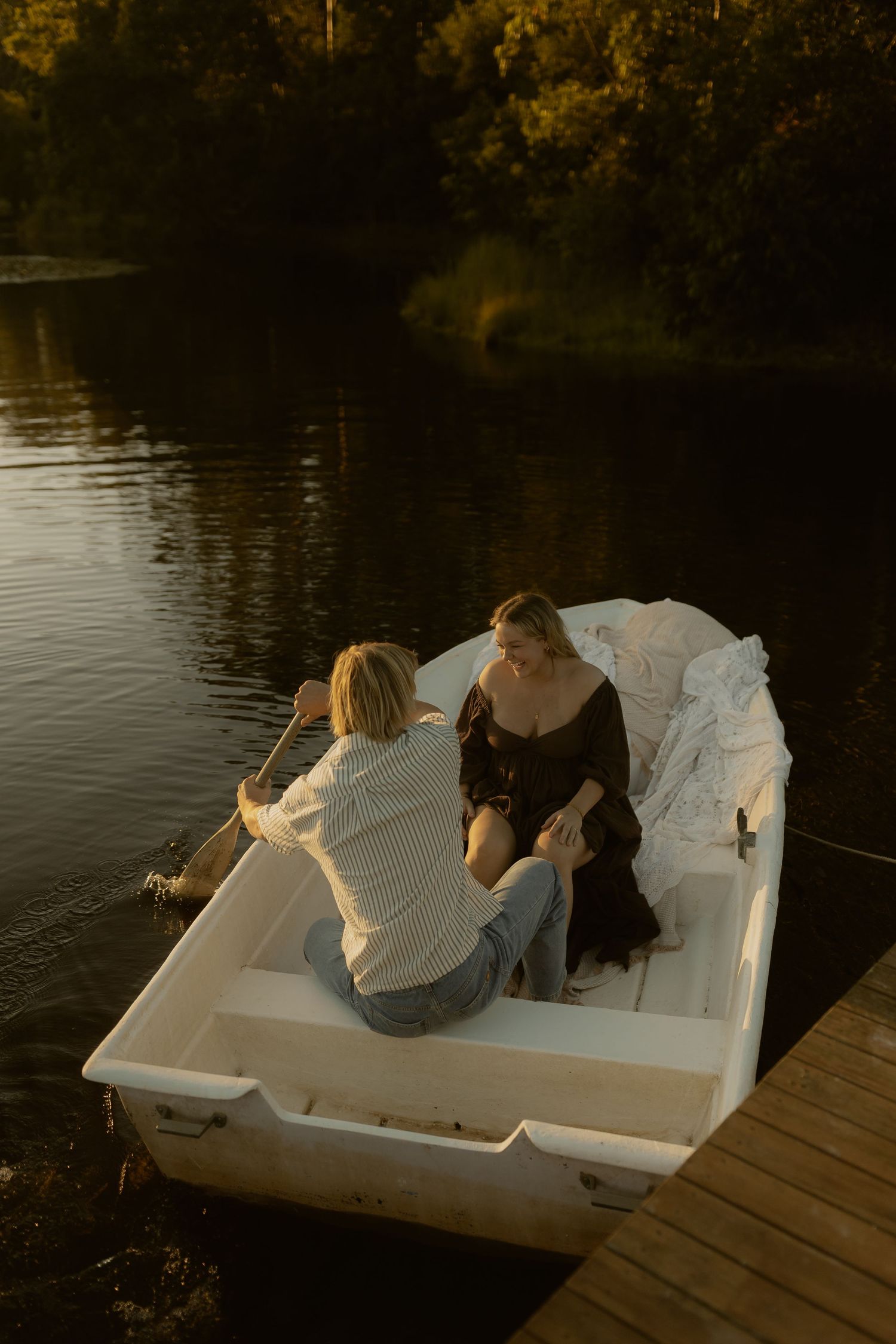 Couple sitting in a white rowboat at golden hour, docked by a wooden pier on a calm forest lake.