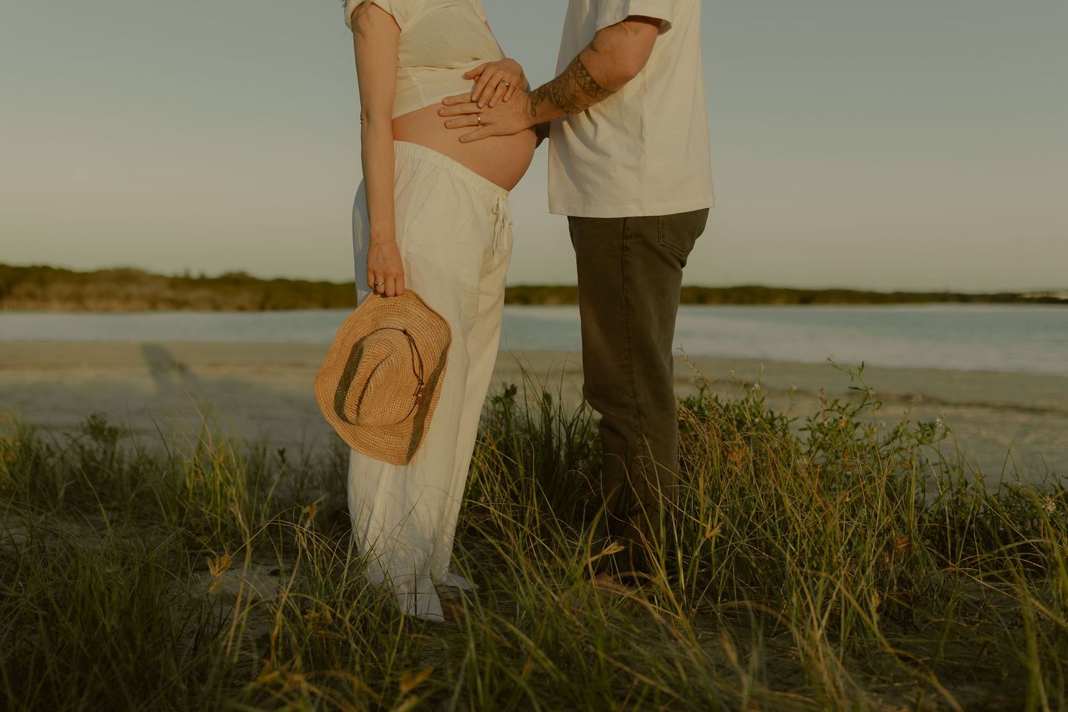 Couple standing close together on a beach at golden hour, woman in white dress holding a straw hat.