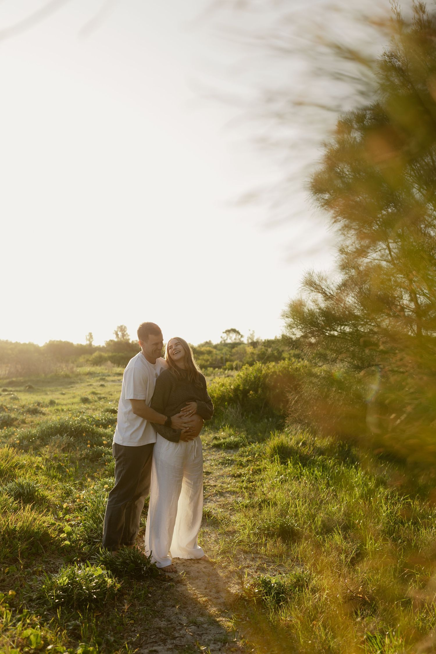 Couple standing together on sunlit nature path, man embracing pregnant partner near pine tree at golden hour.