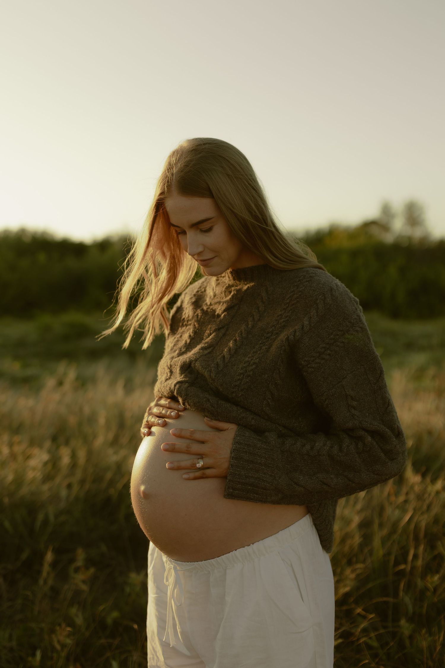Pregnant woman in knit sweater cradling baby bump in golden field at sunset.