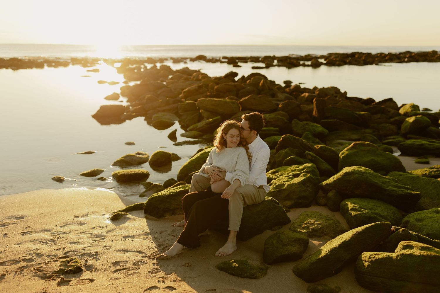 Couple sitting together on mossy rocks at a golden sunset beach, sharing a romantic embrace by the ocean shore.