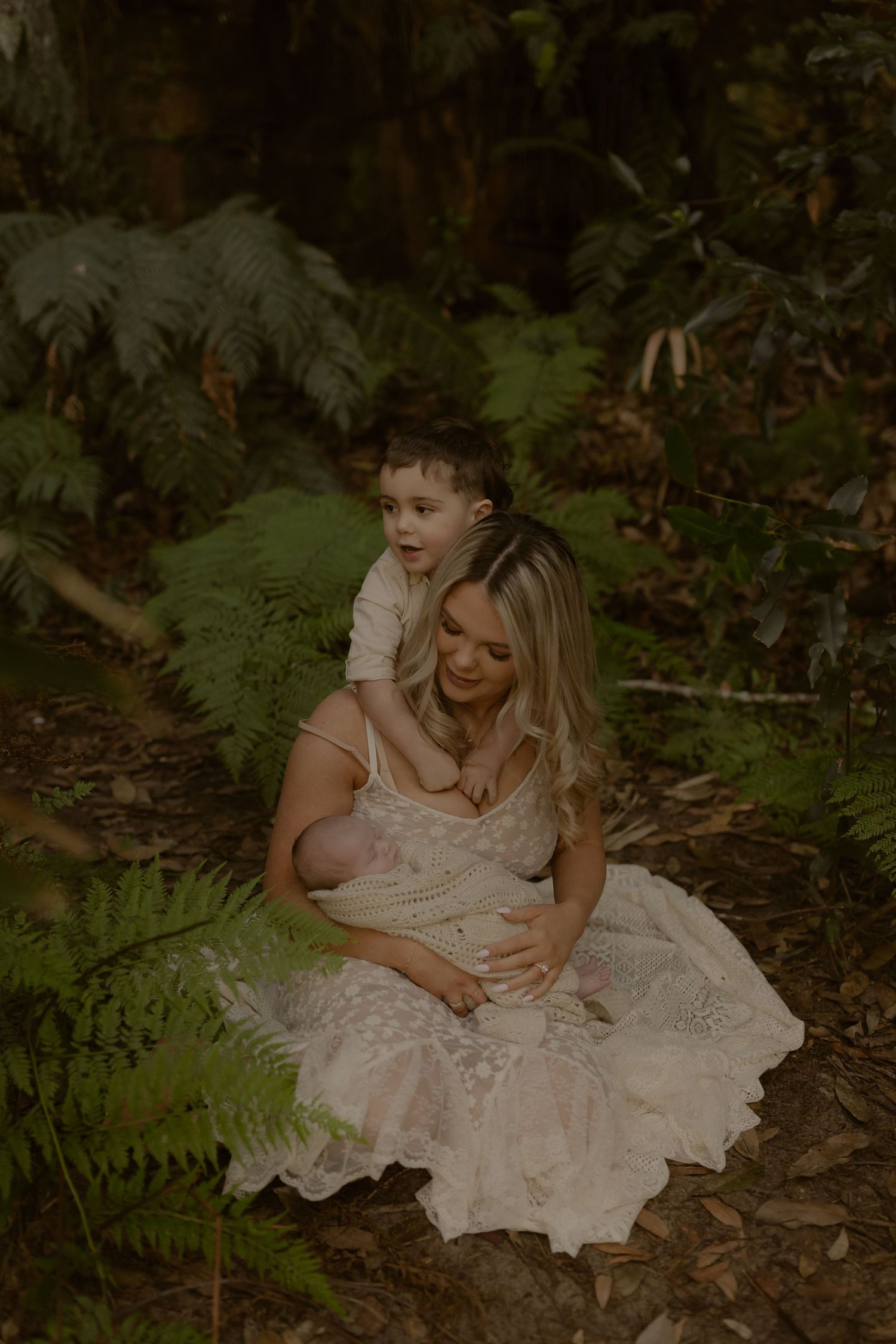 Blonde mother in lace dress sits in fern forest cradling newborn while toddler embraces her from behind.