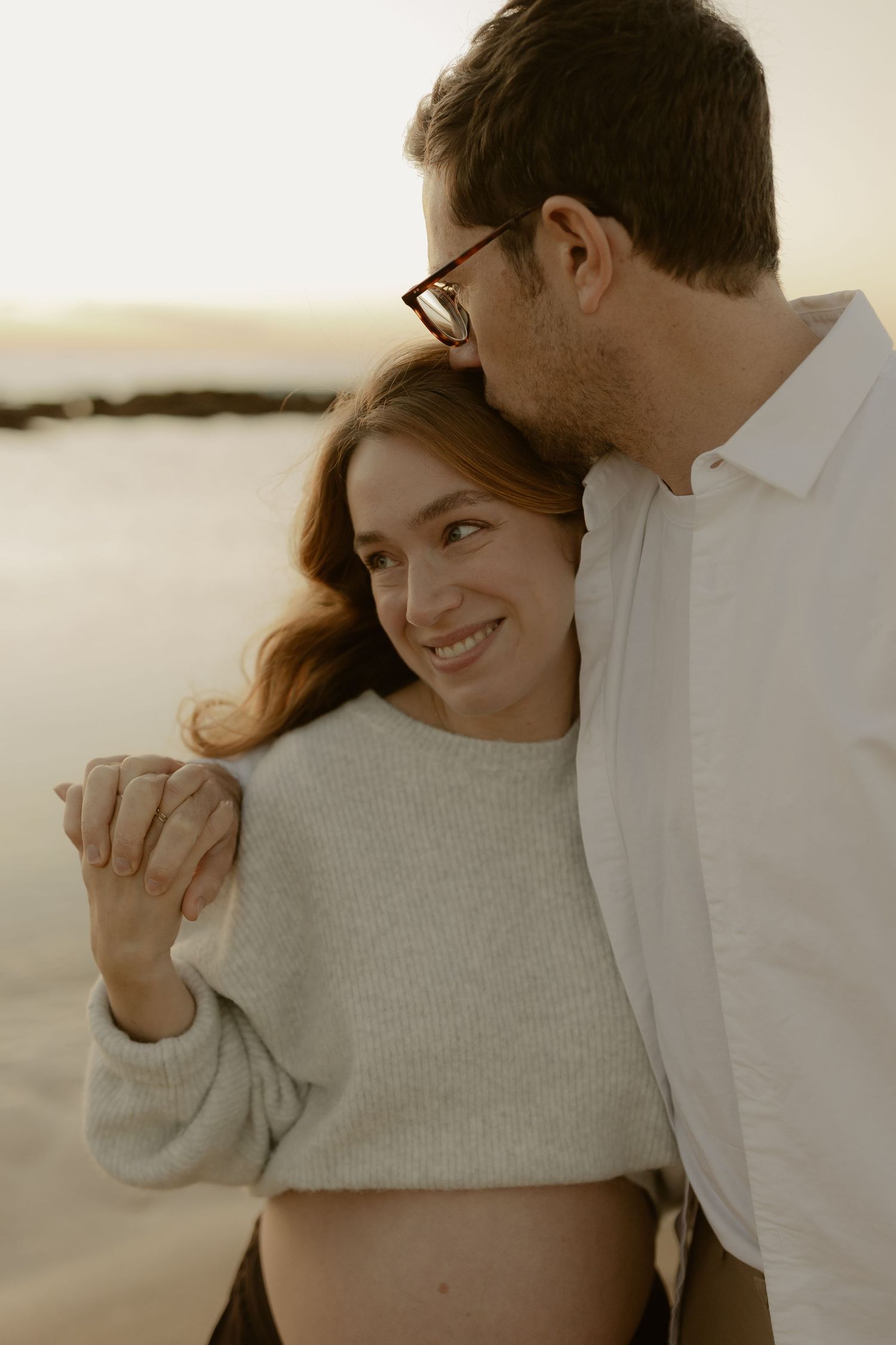 Smiling pregnant woman embraced by man with glasses kissing her head on beach at sunset.