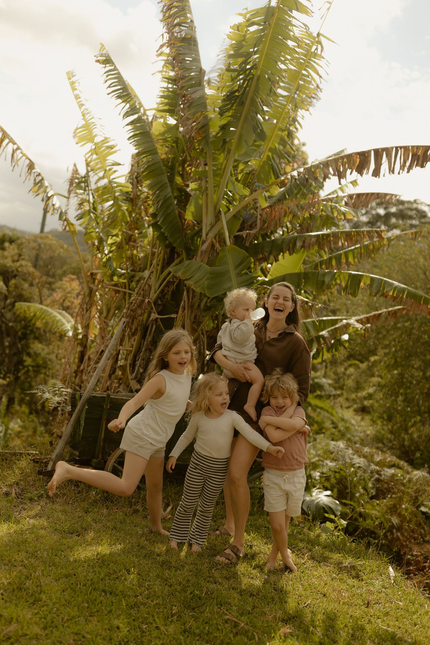 Joyful mother holding toddler surrounded by three children near tropical banana tree outdoors.