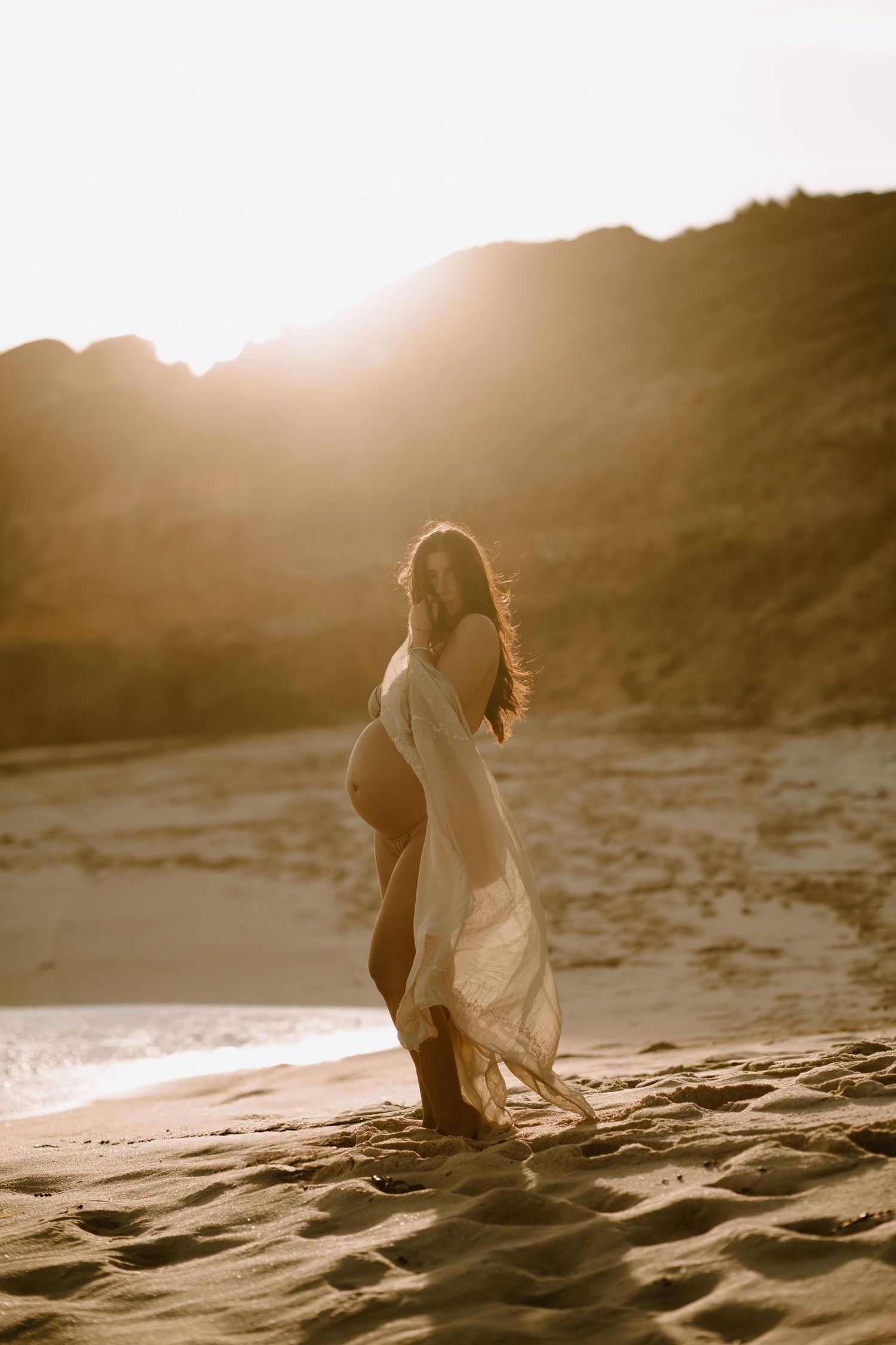 A silhouette stands on a sandy beach at sunset wearing a flowing white dress as golden light filters through the hills.