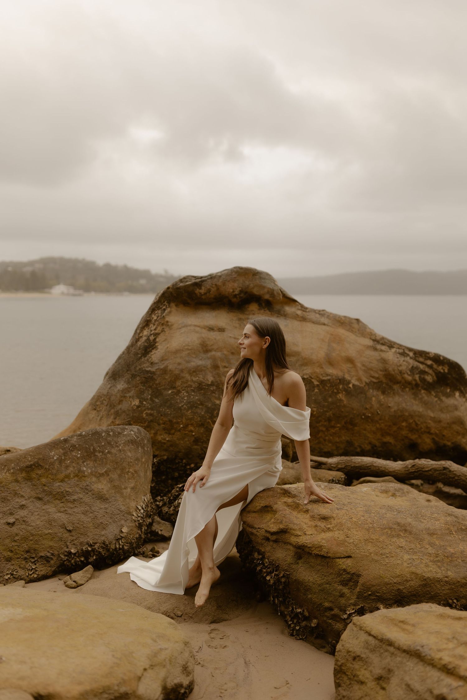 A person in a flowing white dress sits gracefully on coastal rocks with the ocean in the background.