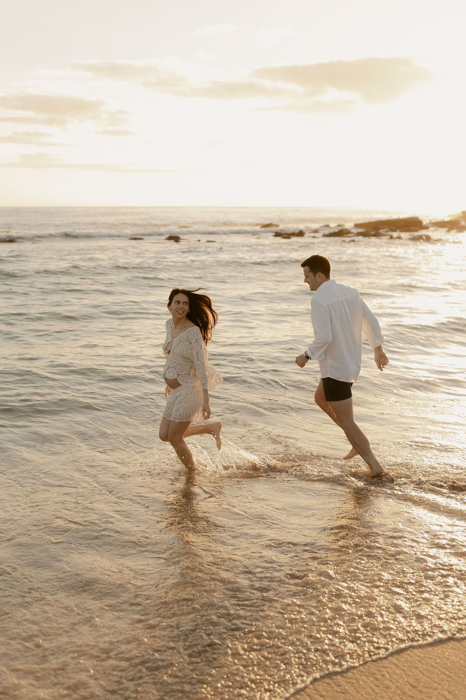 A couple playfully runs and splashes in the ocean waves at sunset along a golden sandy beach.