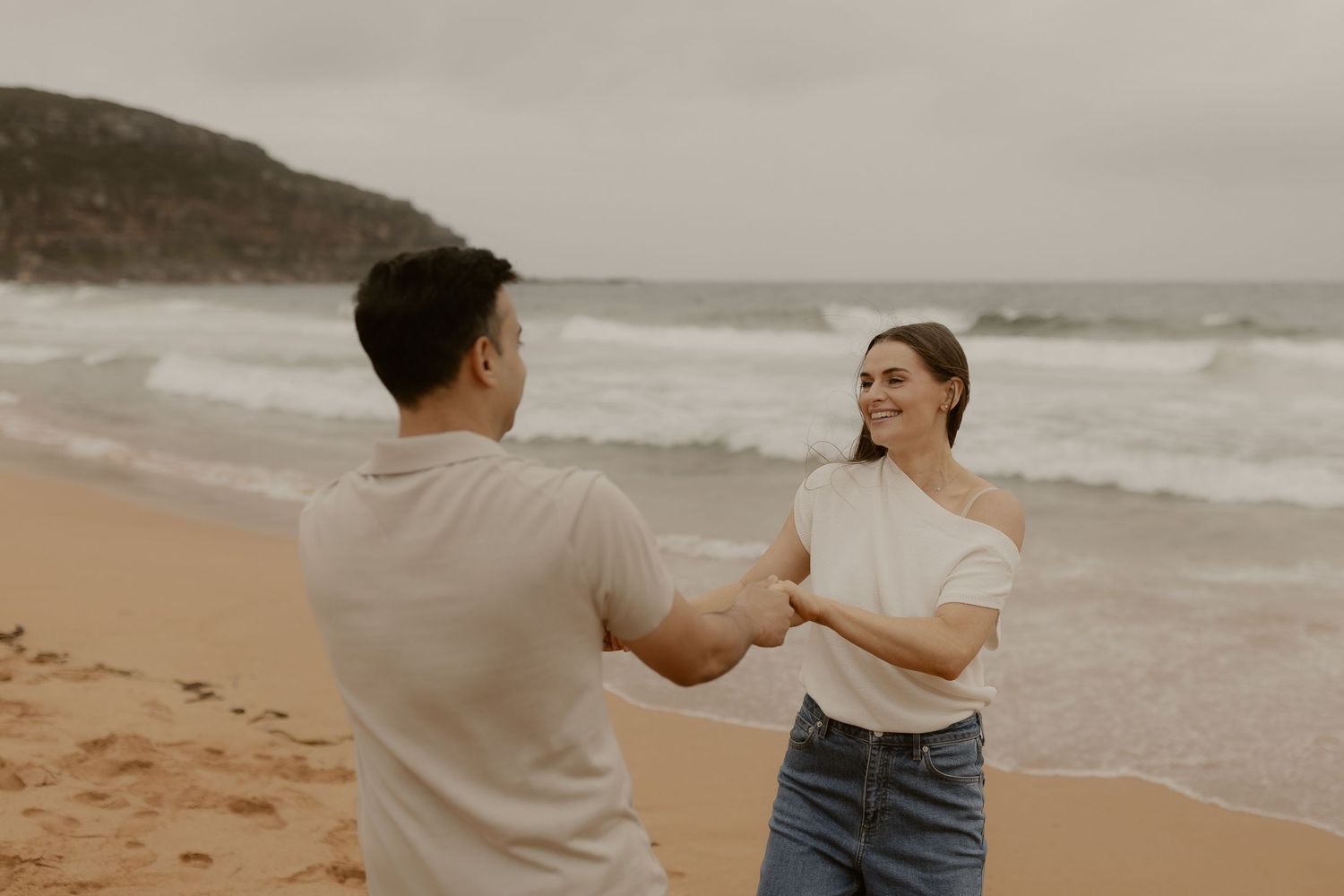 Couple in casual attire dances together on a sandy beach at sunset with ocean waves in the background.