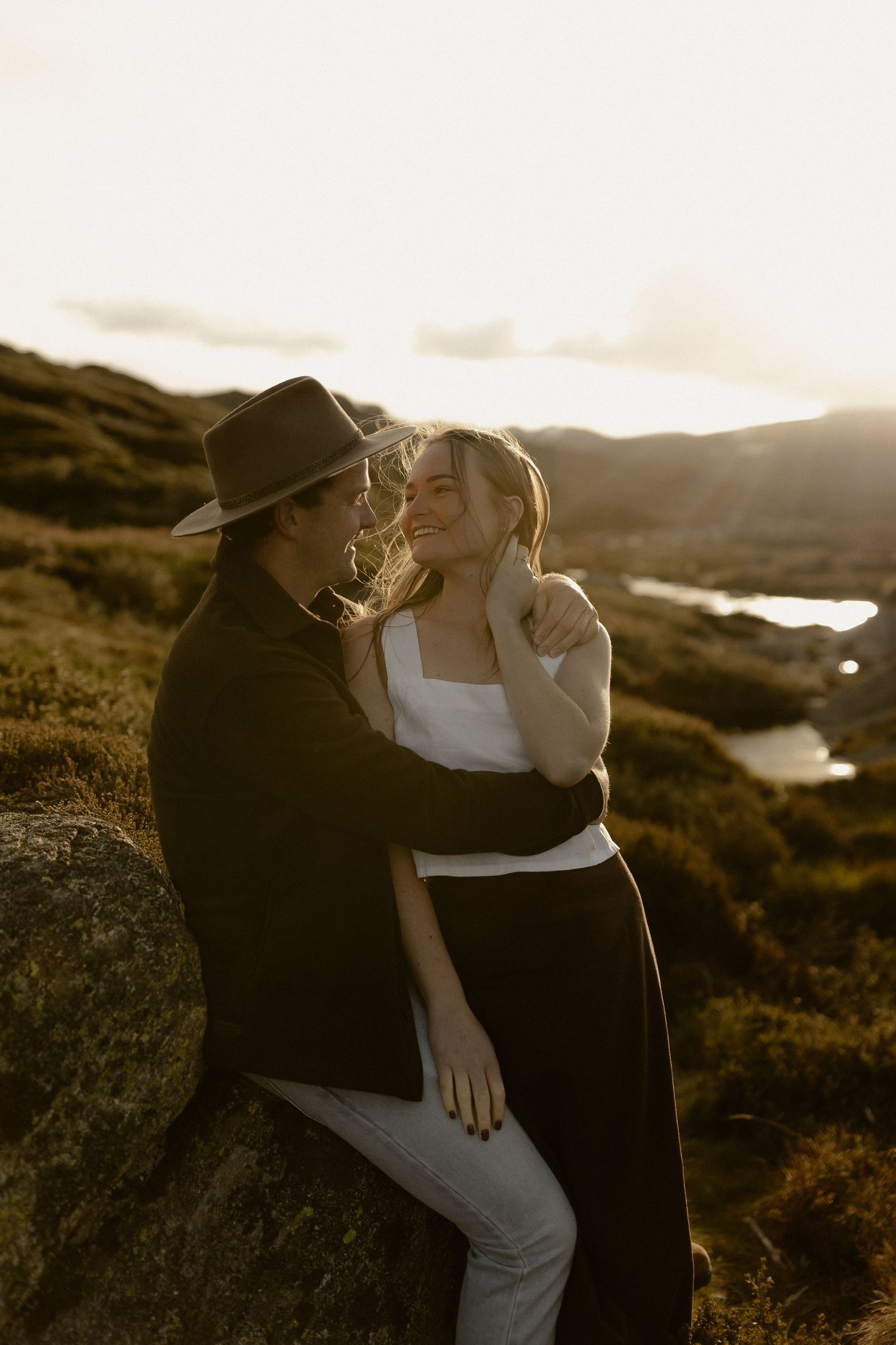 A couple embraces while standing on rocky terrain with mountains and water in the distance at sunset.