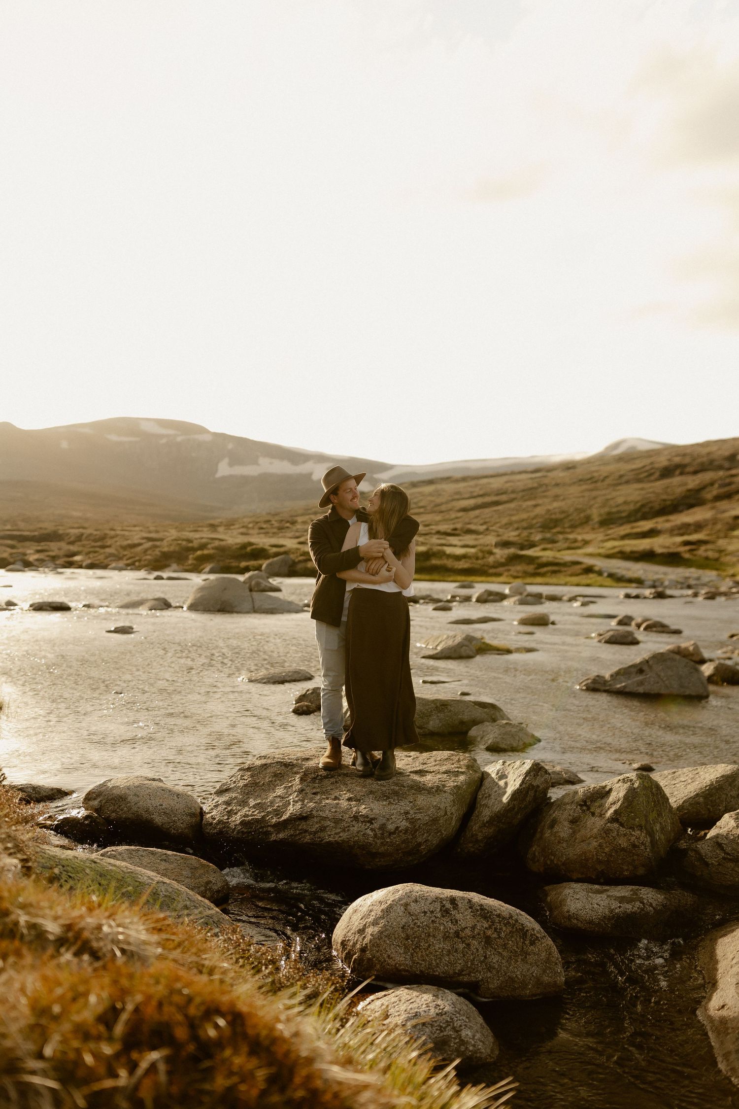 Couple sitting on coastal rocks at sunset with mountains and water in the background.