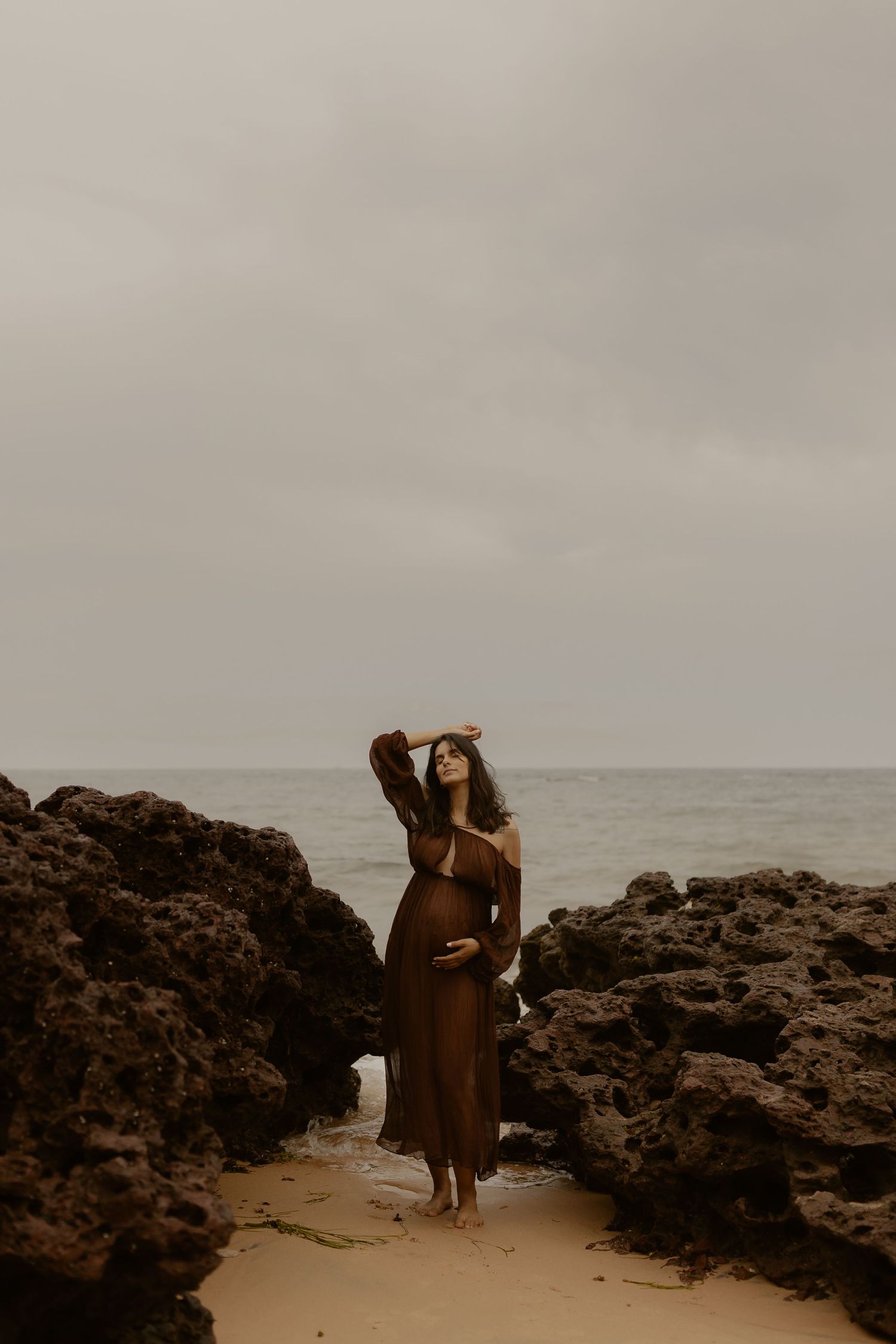 A person in a flowing brown dress poses gracefully on a rocky beach at sunset.