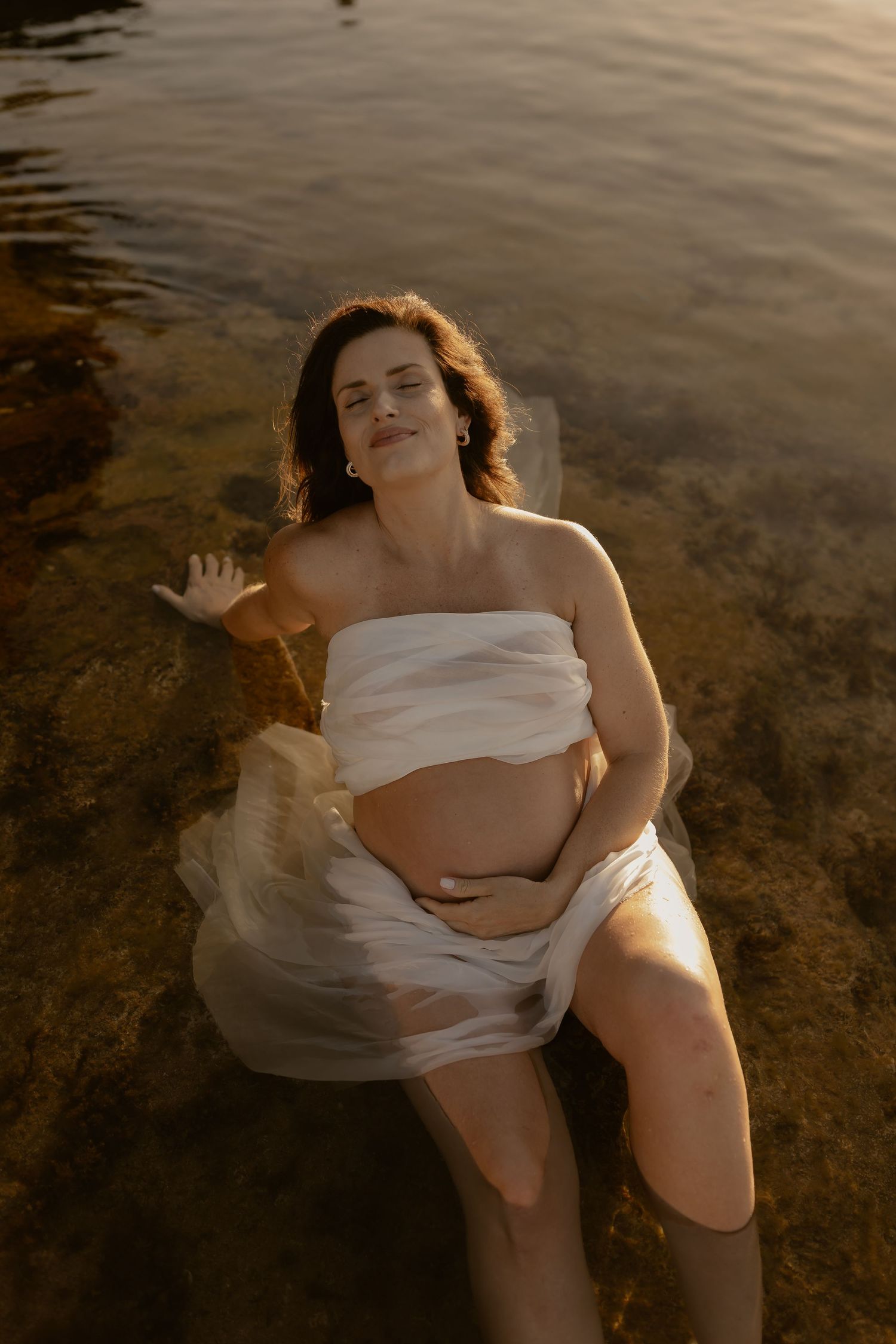 Ethereal maternity photo of a woman in white fabric floating in golden-lit water during sunset.