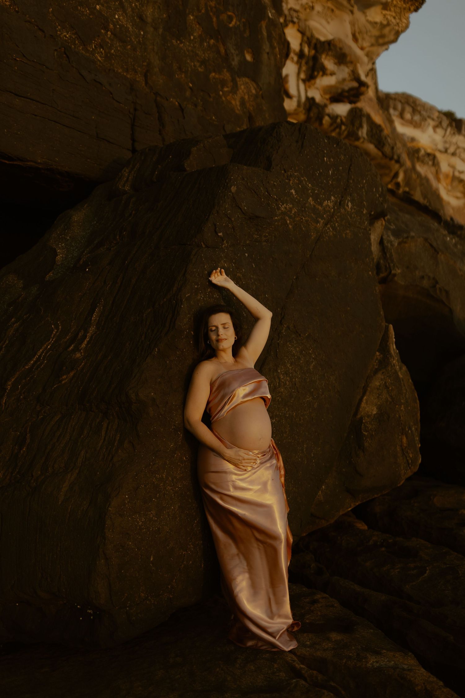 A person poses gracefully against a dark rocky cliff face in warm evening light.