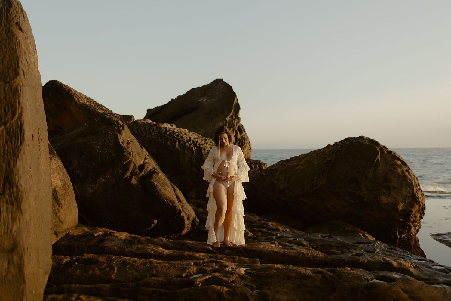 A couple stands on rocky coastal cliffs overlooking the ocean at sunset during their engagement photoshoot.