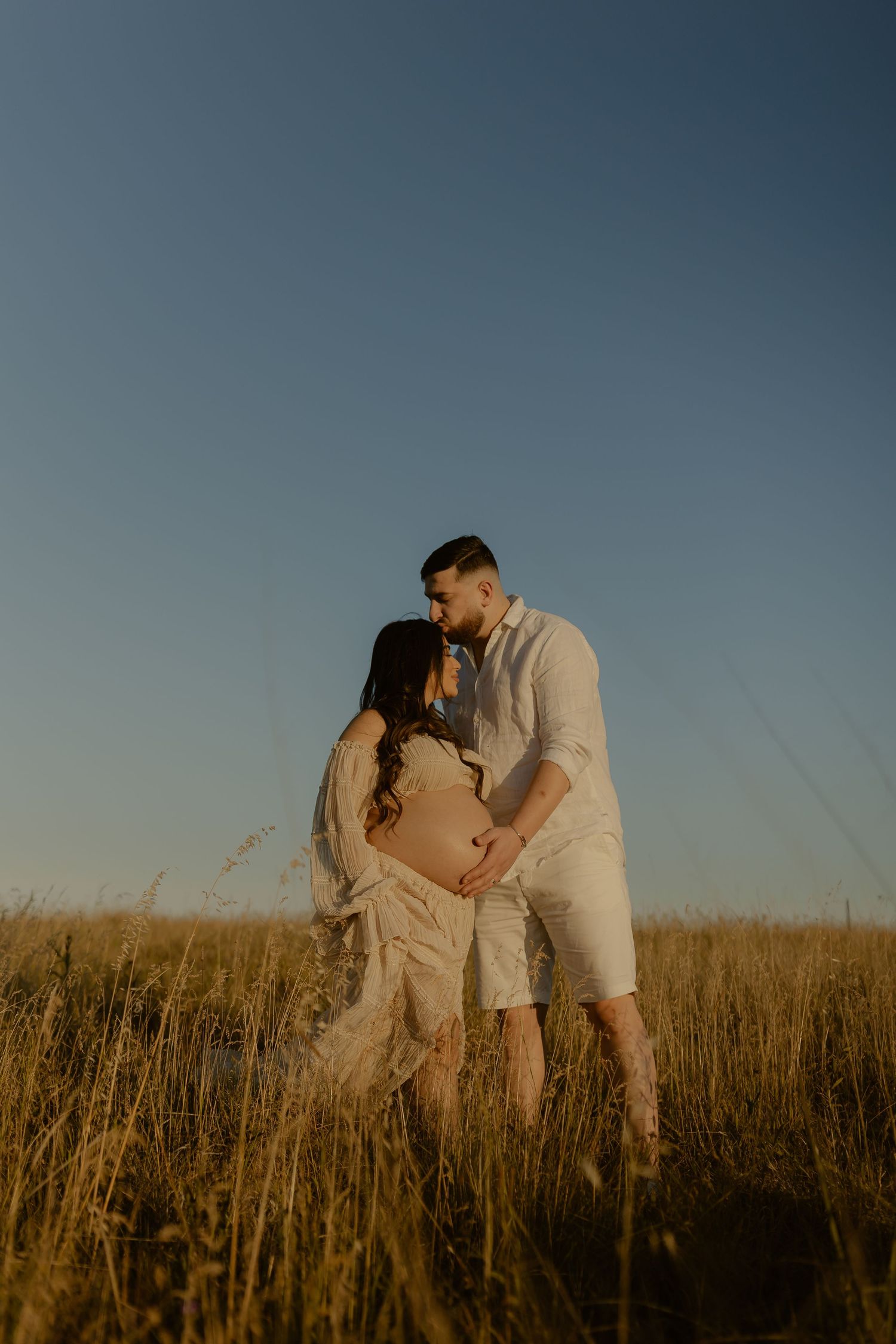 A romantic couple in white outfits embraces in a golden wheat field at sunset.