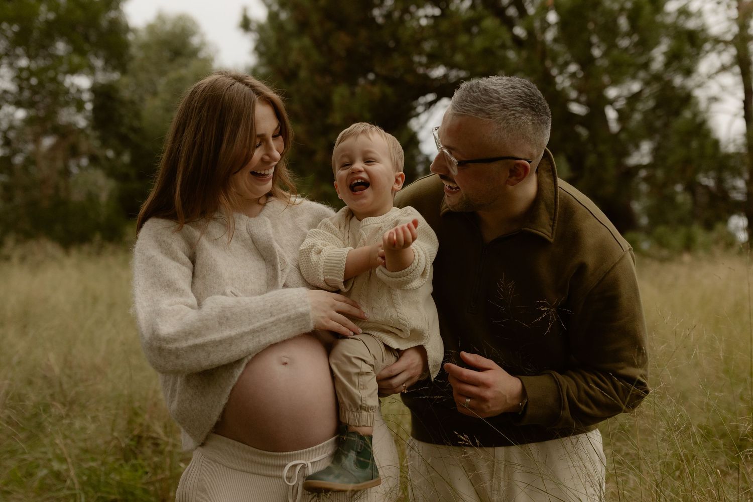 Pregnant woman and man laughing with toddler in a grassy outdoor setting during a maternity photo session.