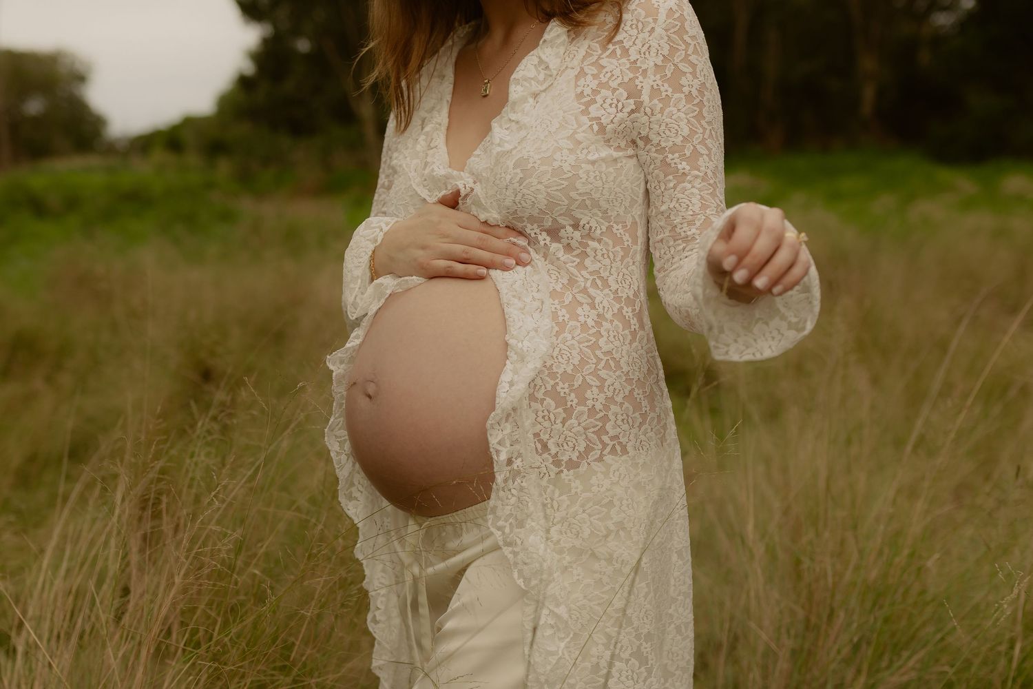 Close-up of pregnant woman in white lace dress cradling baby bump outdoors in natural setting.