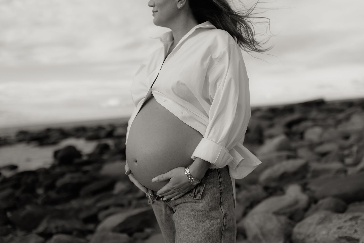 Black and white photo of a pregnant woman in an open white shirt showing her baby bump on a beach.
