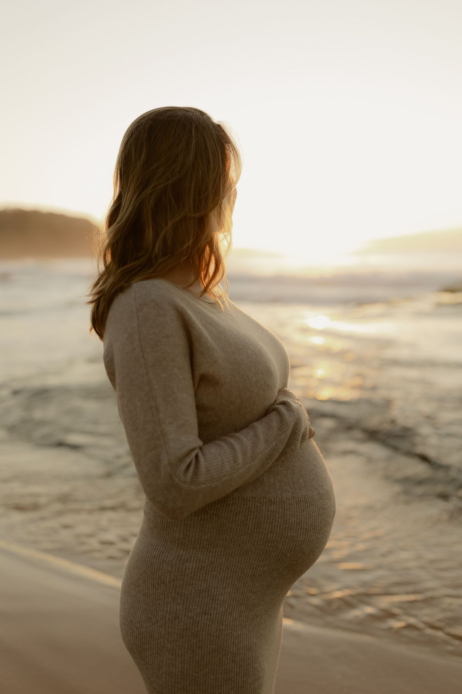 Pregnant woman in beige knit dress holding baby bump at golden hour beach sunset.