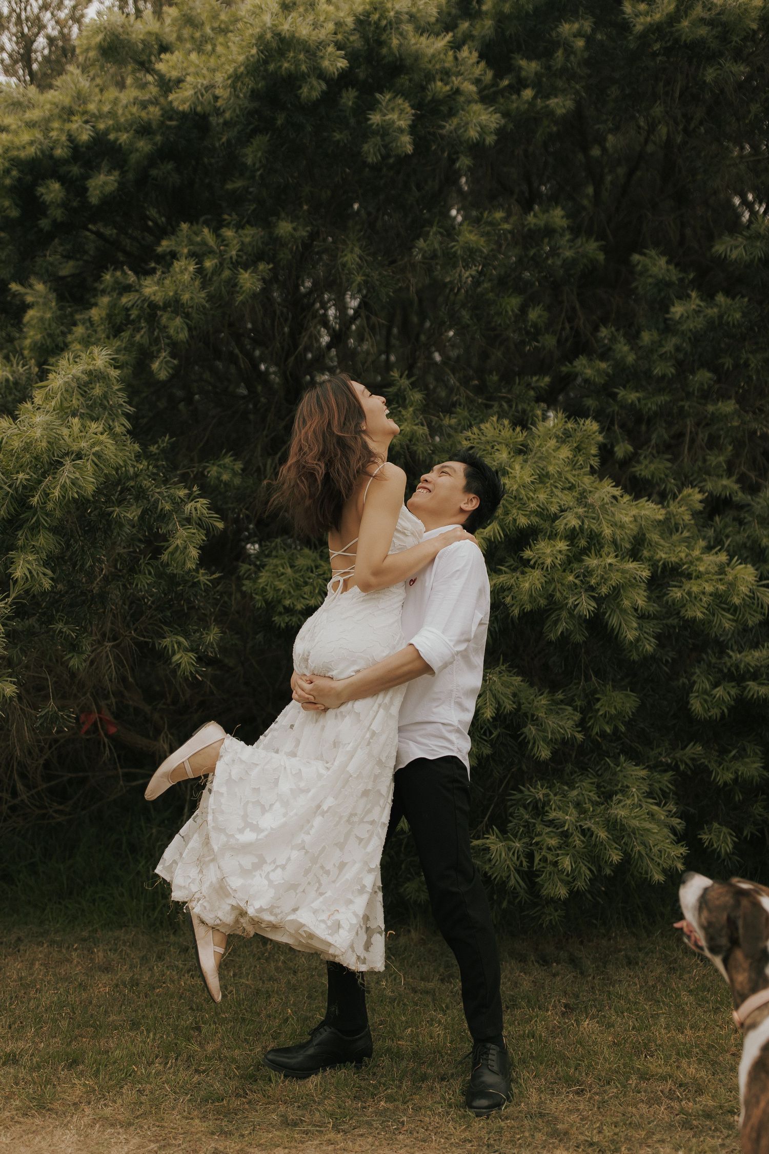 A couple dances and twirls together on grass with trees in the background while a dog watches nearby.