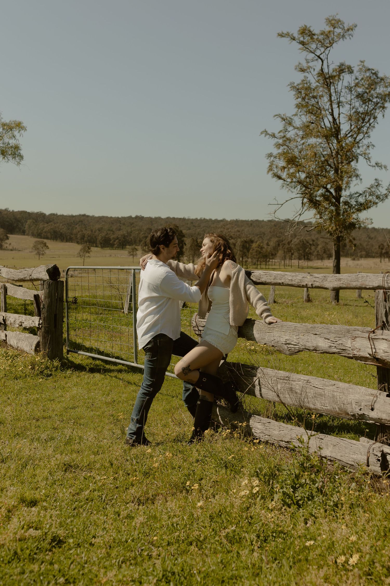 Couple playfully poses by a wooden fence in a rustic countryside setting.