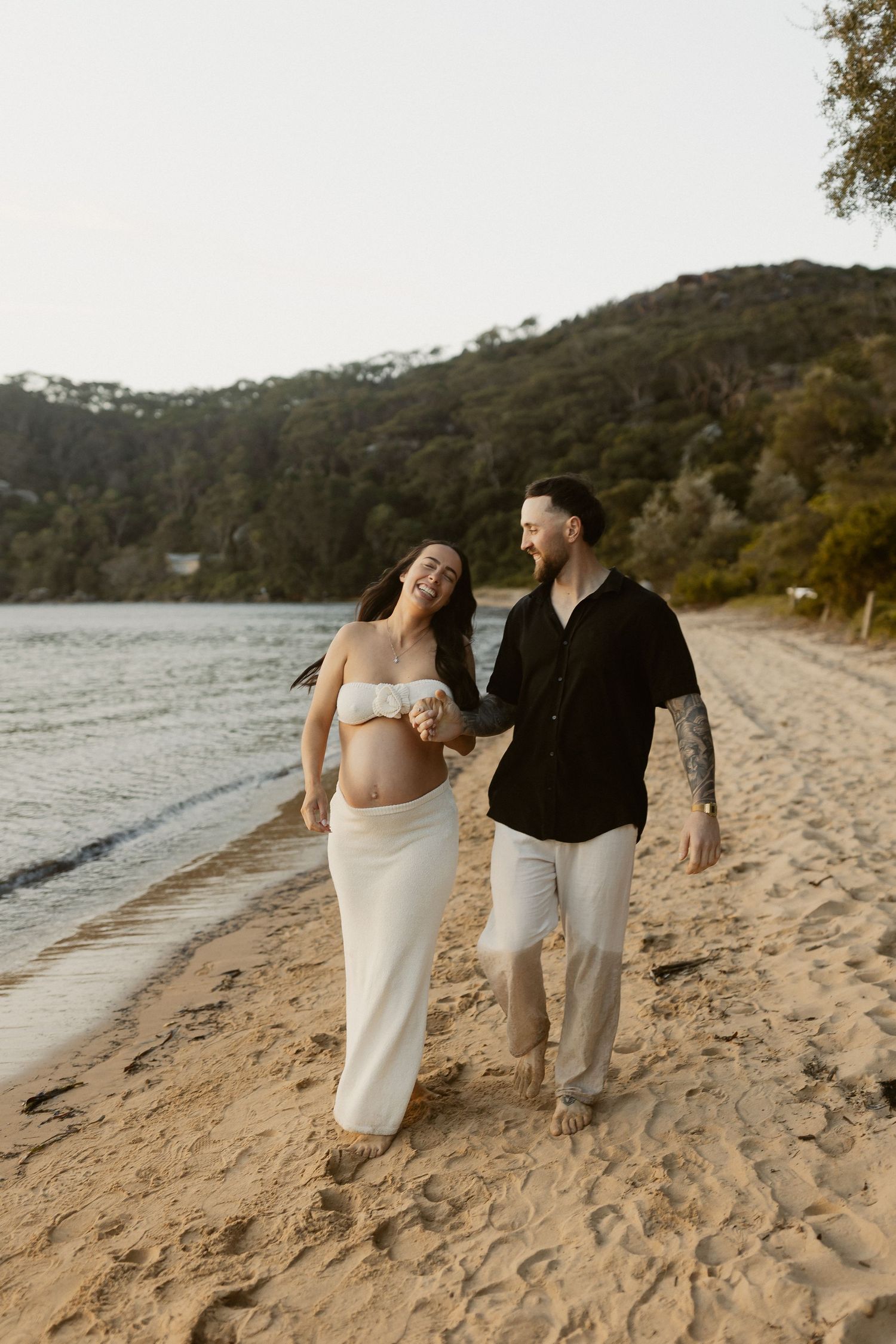 A couple walks hand in hand along a secluded beach with hills in the background during sunset.