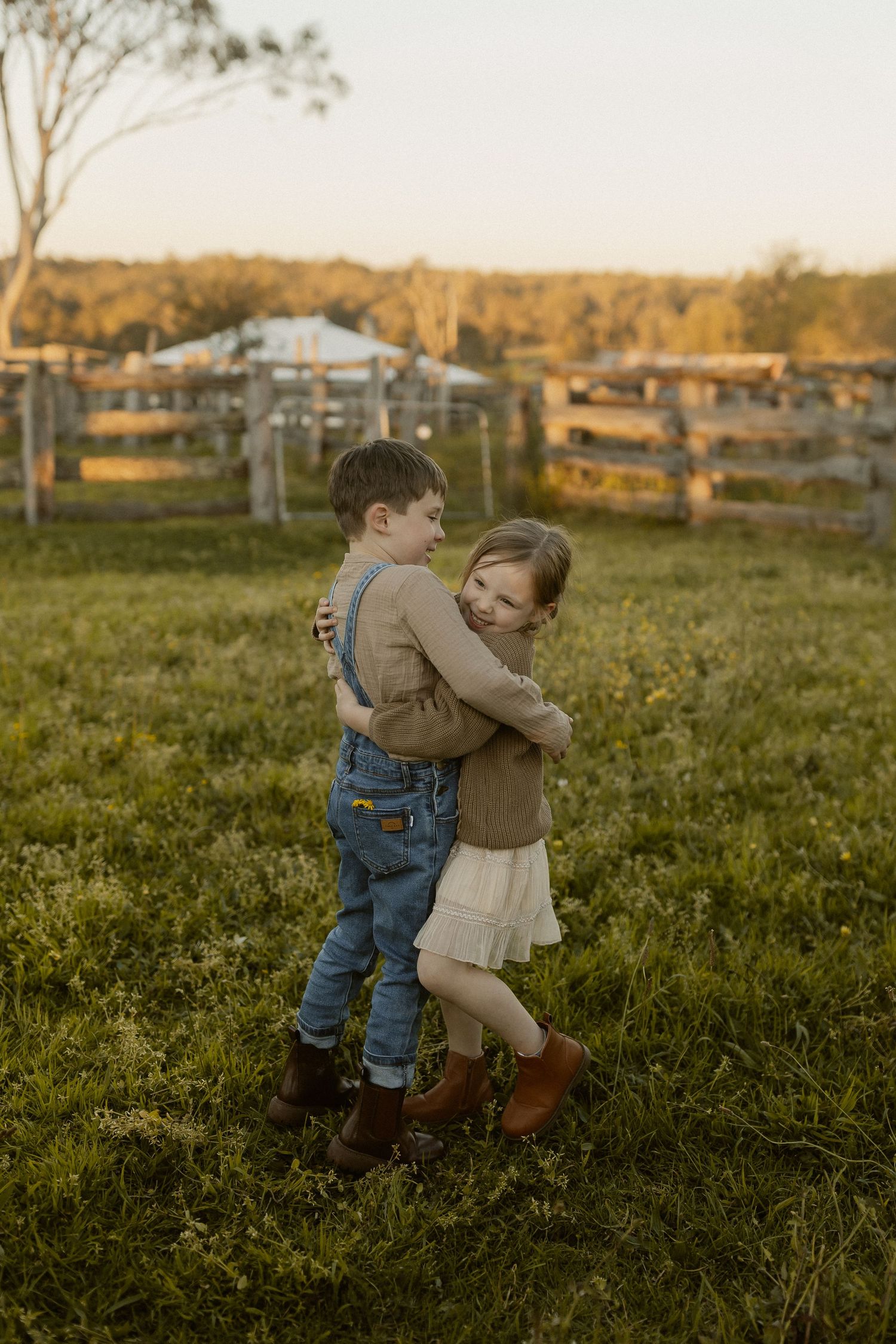 Two young children dancing together in a rustic field during golden hour with farm buildings in background.