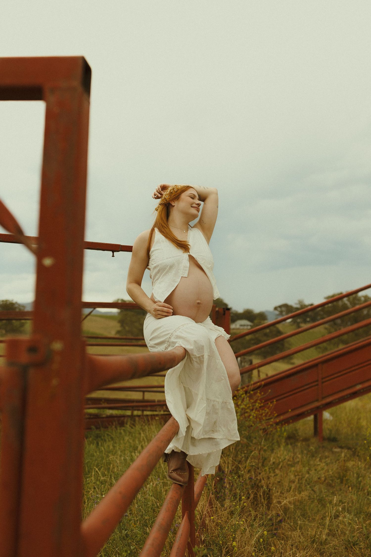 Figure in a white dress poses gracefully against rustic metal structures in a serene outdoor setting at sunset.