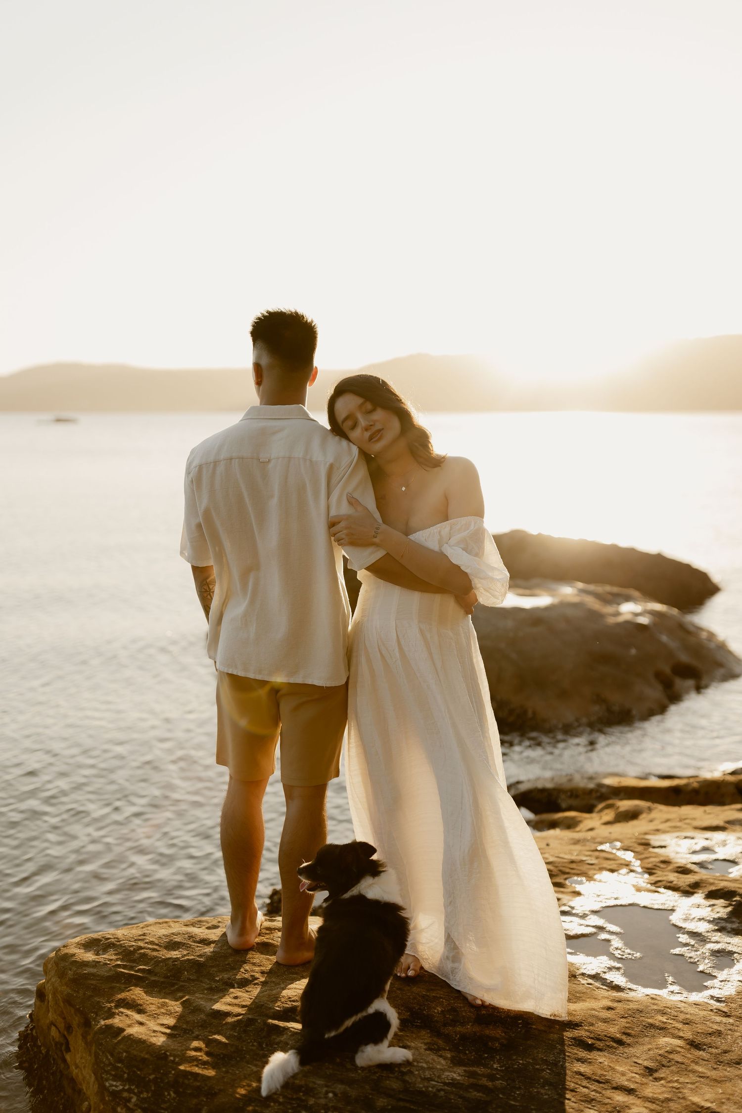 A couple stands together at sunset on rocky coastal shores with their dog beside them.
